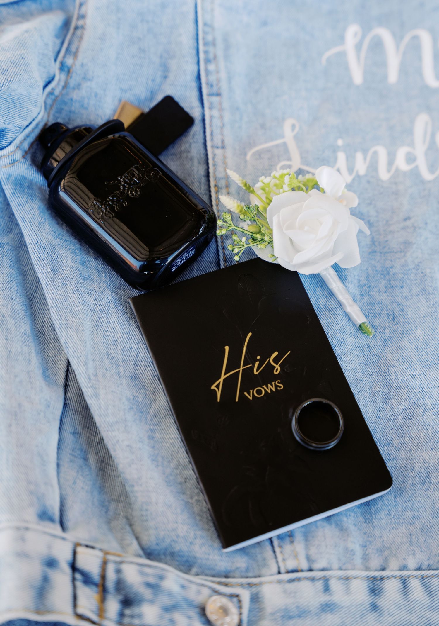 His Vows black book with gold text, cologne bottle, boutonniere, and ring on a denim jacket.
