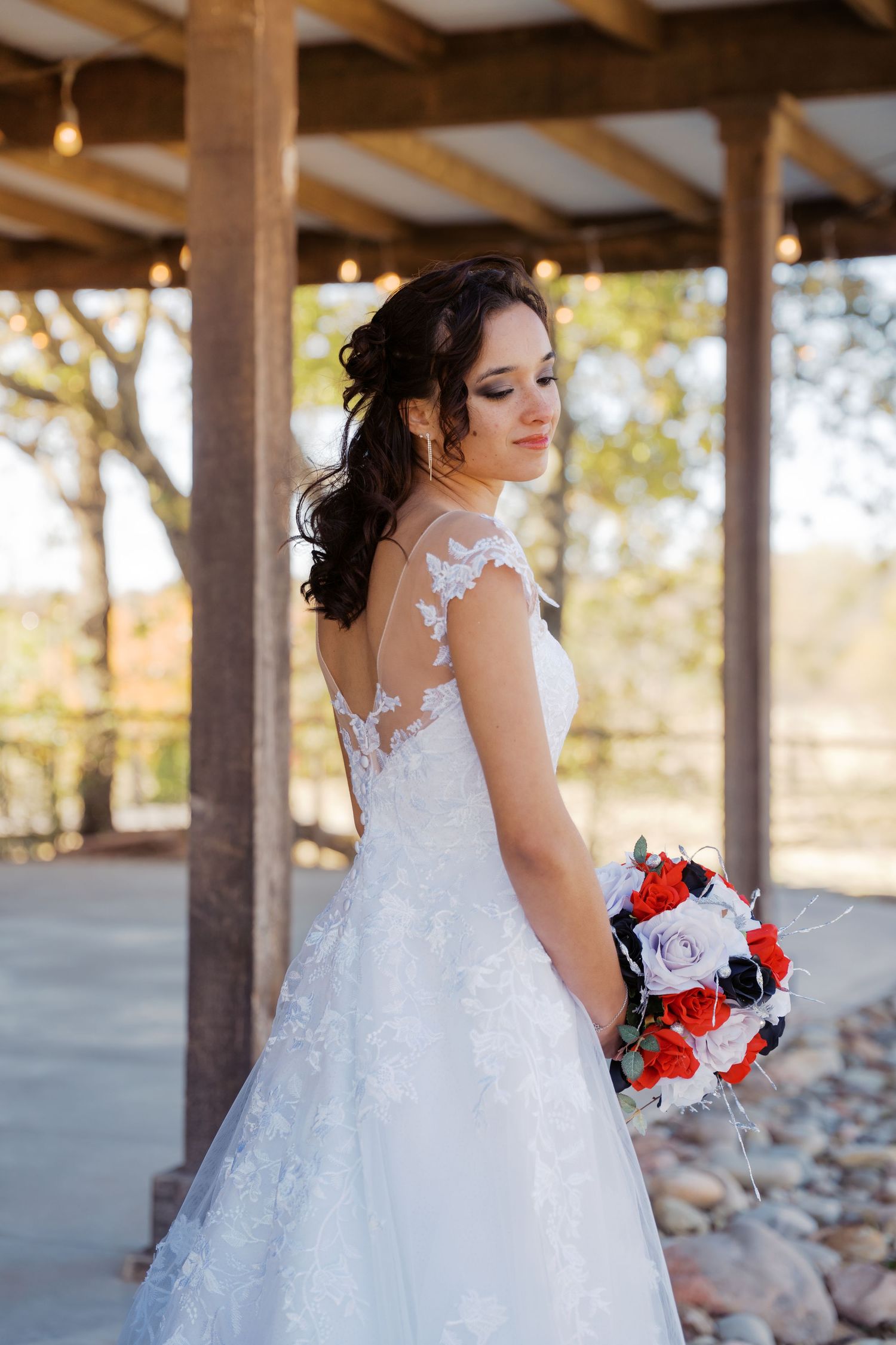 Bride in lace wedding gown holds red and black bouquet under wooden pergola, looking over shoulder. Oklahoma wedding photographer.