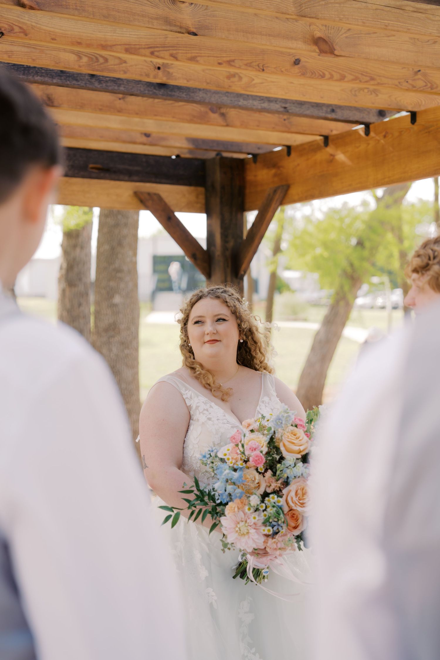 Bride holding colorful pastel bouquet of flowers during outdoor wedding ceremony under wooden pergola.