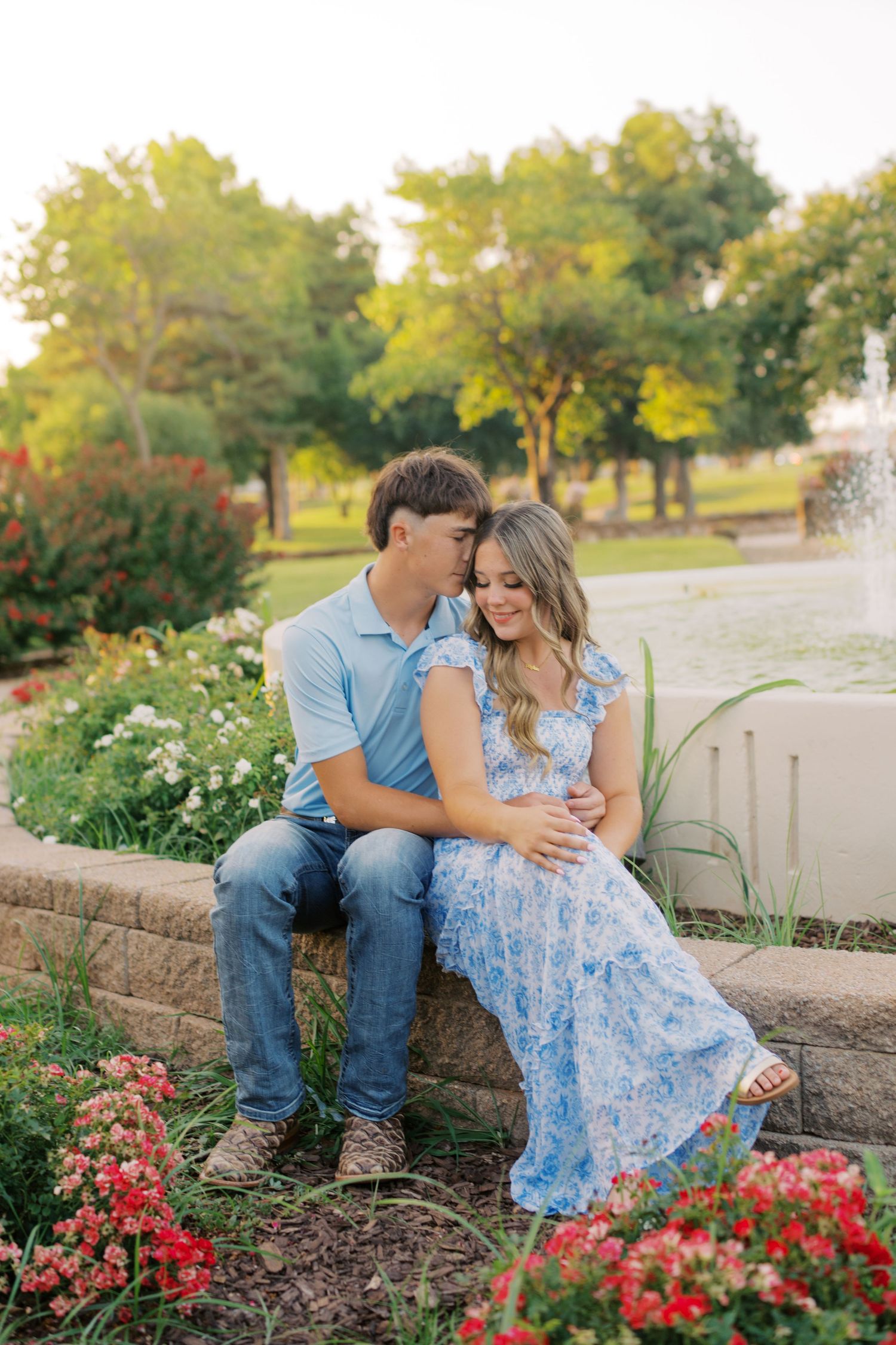 Couple sitting together by a fountain surrounded by colorful flowers in a lush park at golden hour.
