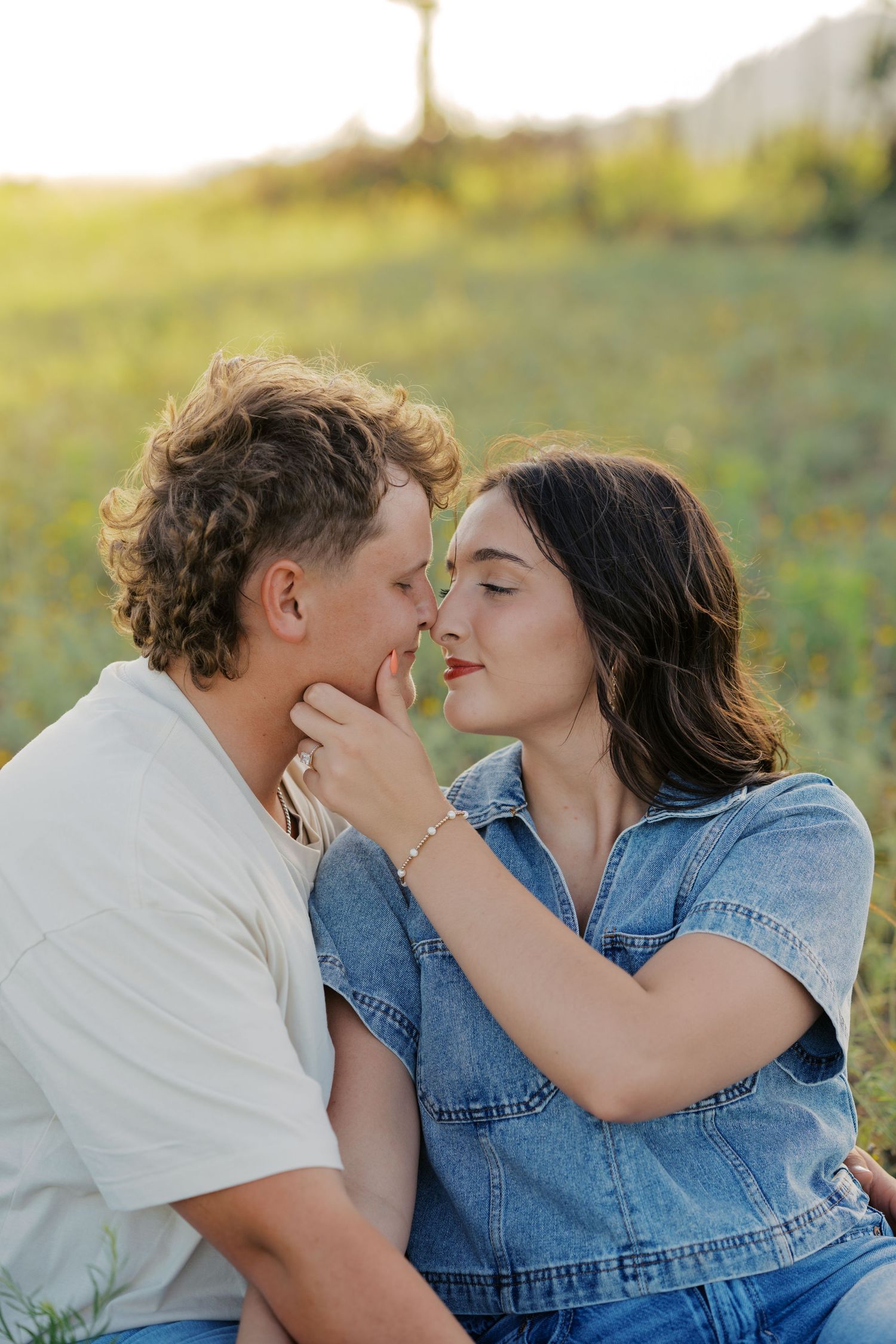 Romantic couple about to kiss while sitting in a sunny green wildflower field at sunset.