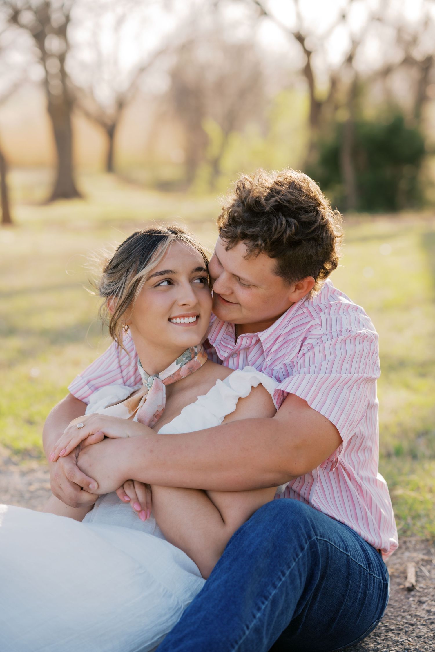 Young engaged couple sitting outdoors, smiling and embracing in soft spring evening light.