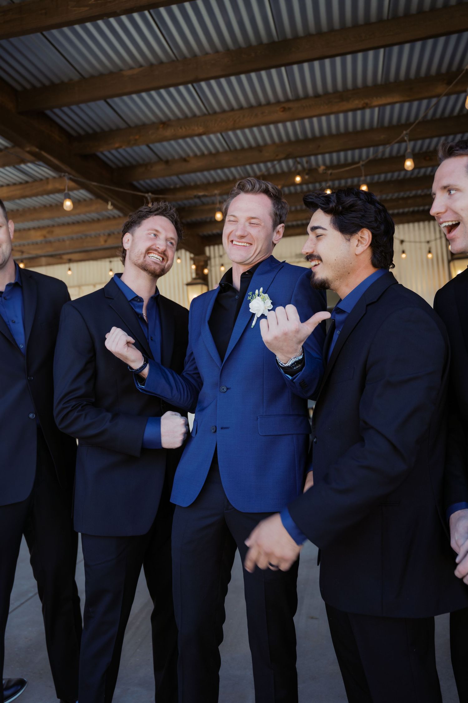 Groom in blue suit with boutonniere laughs with groomsmen in black suits at rustic wedding venue. Oklahoma wedding photographer.