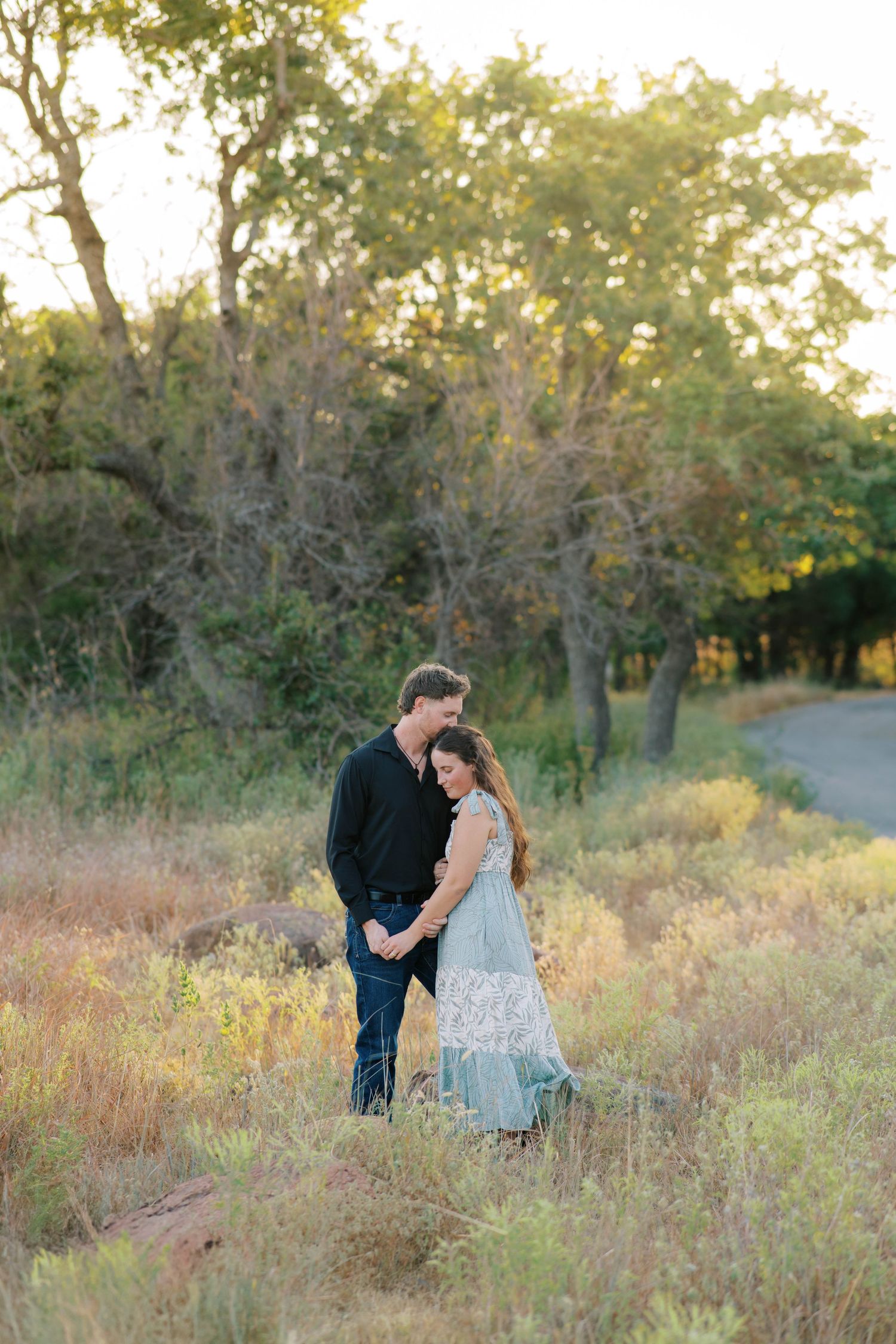 A couple embraces tenderly in a golden sunlit meadow surrounded by wild grass and trees.