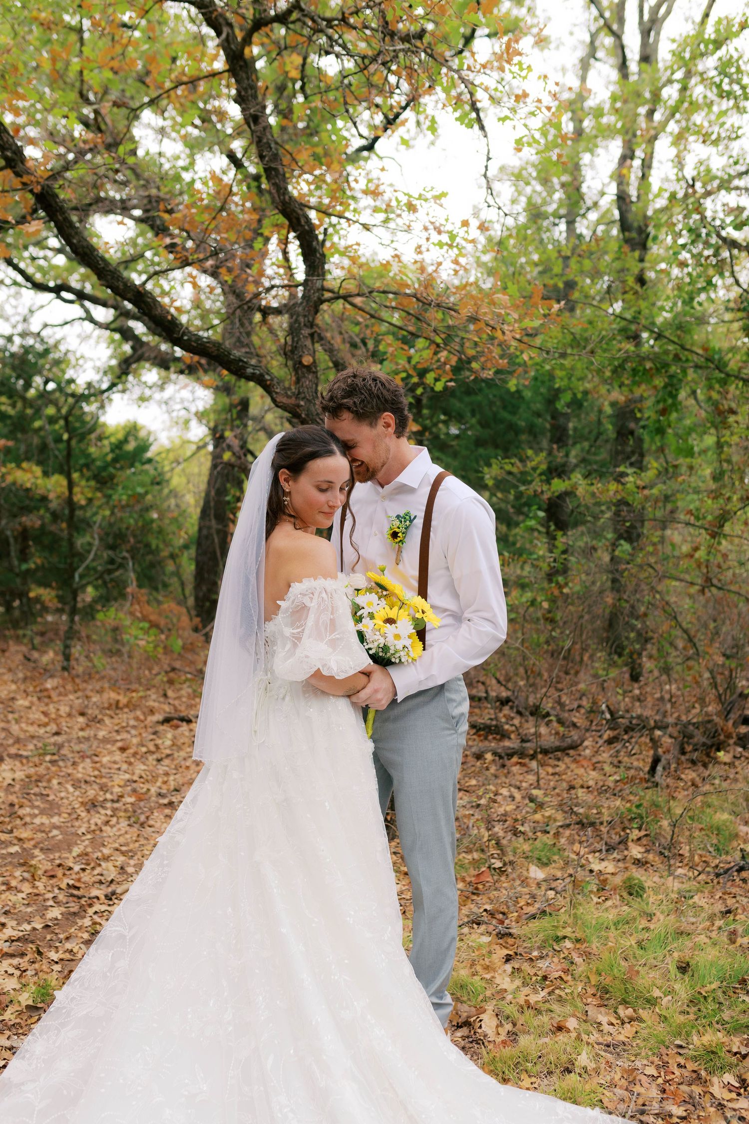 Bride and groom embracing in autumn forest, bride holding yellow sunflower bouquet in white off-shoulder gown with veil. Oklahoma wedding.