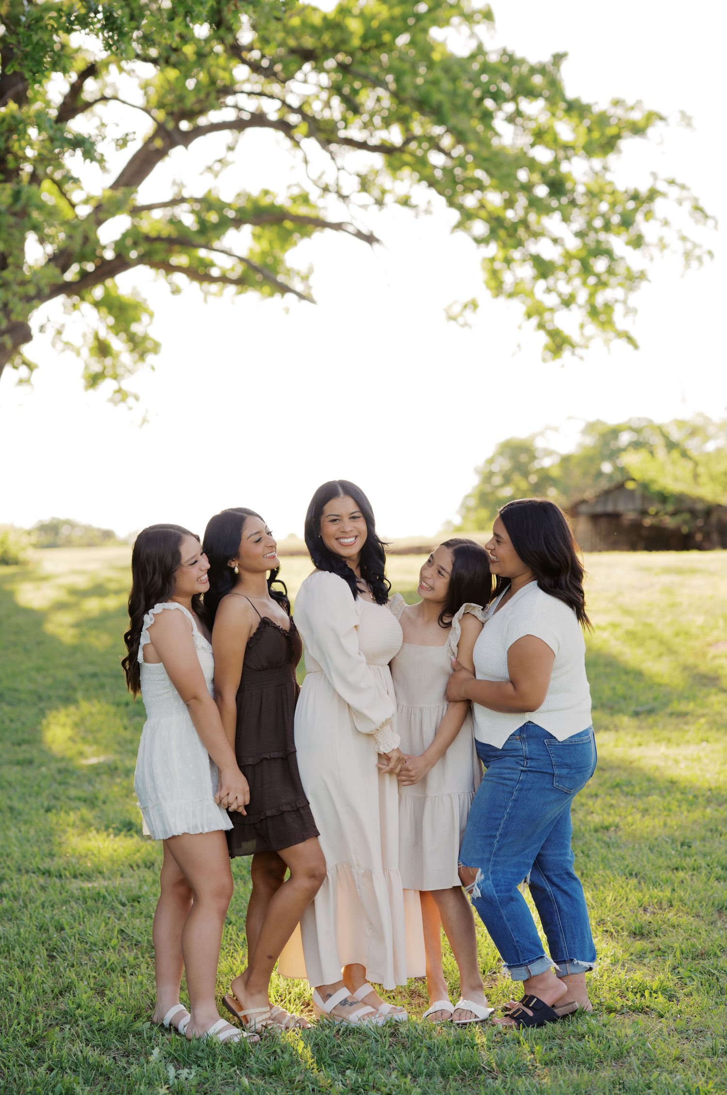 Five women posing together outdoors on green grass under a large tree during golden hour in a family portrait.