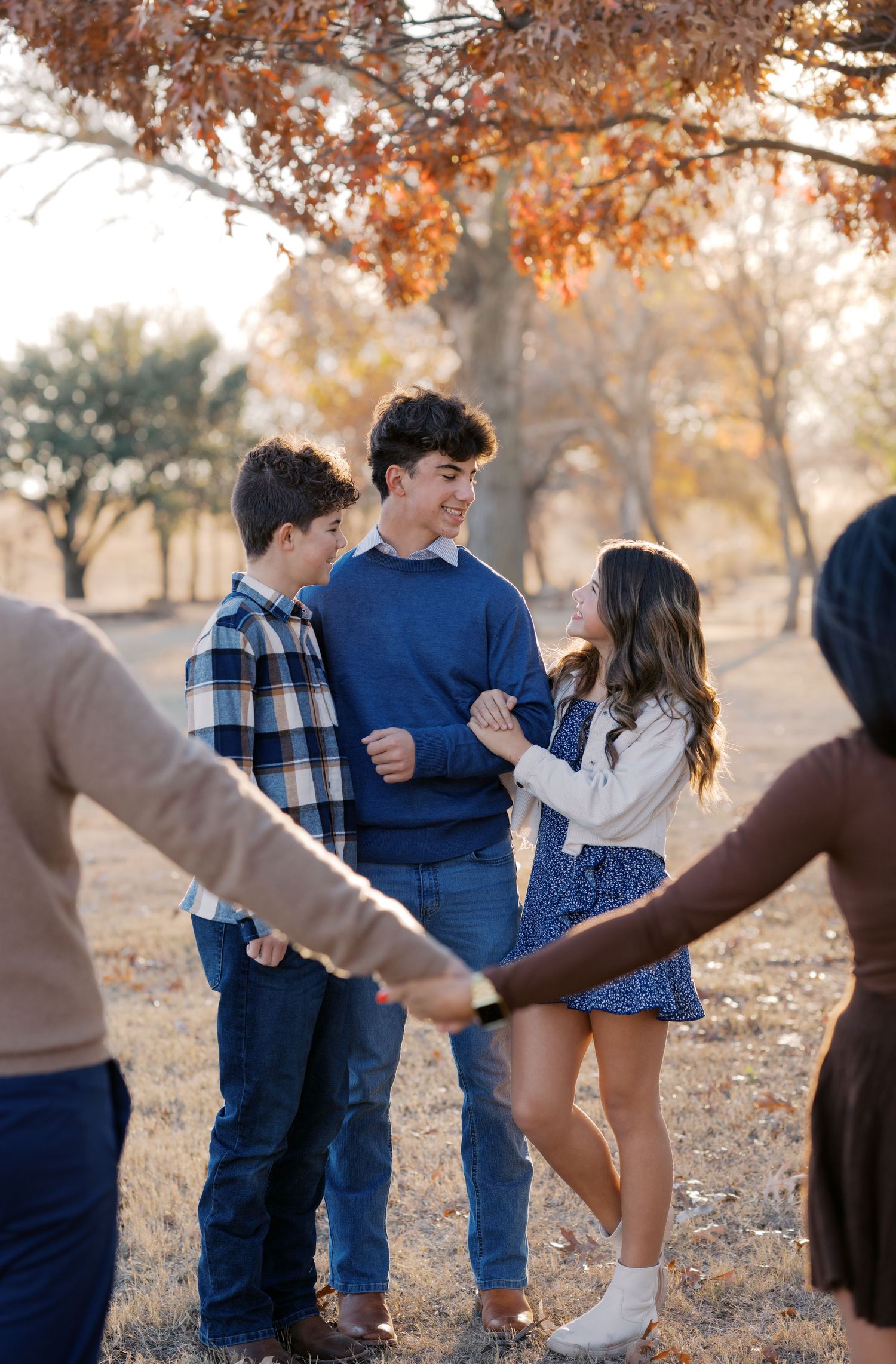 Family holding hands in a circle outdoors during fall, teens laughing together under autumn trees.
