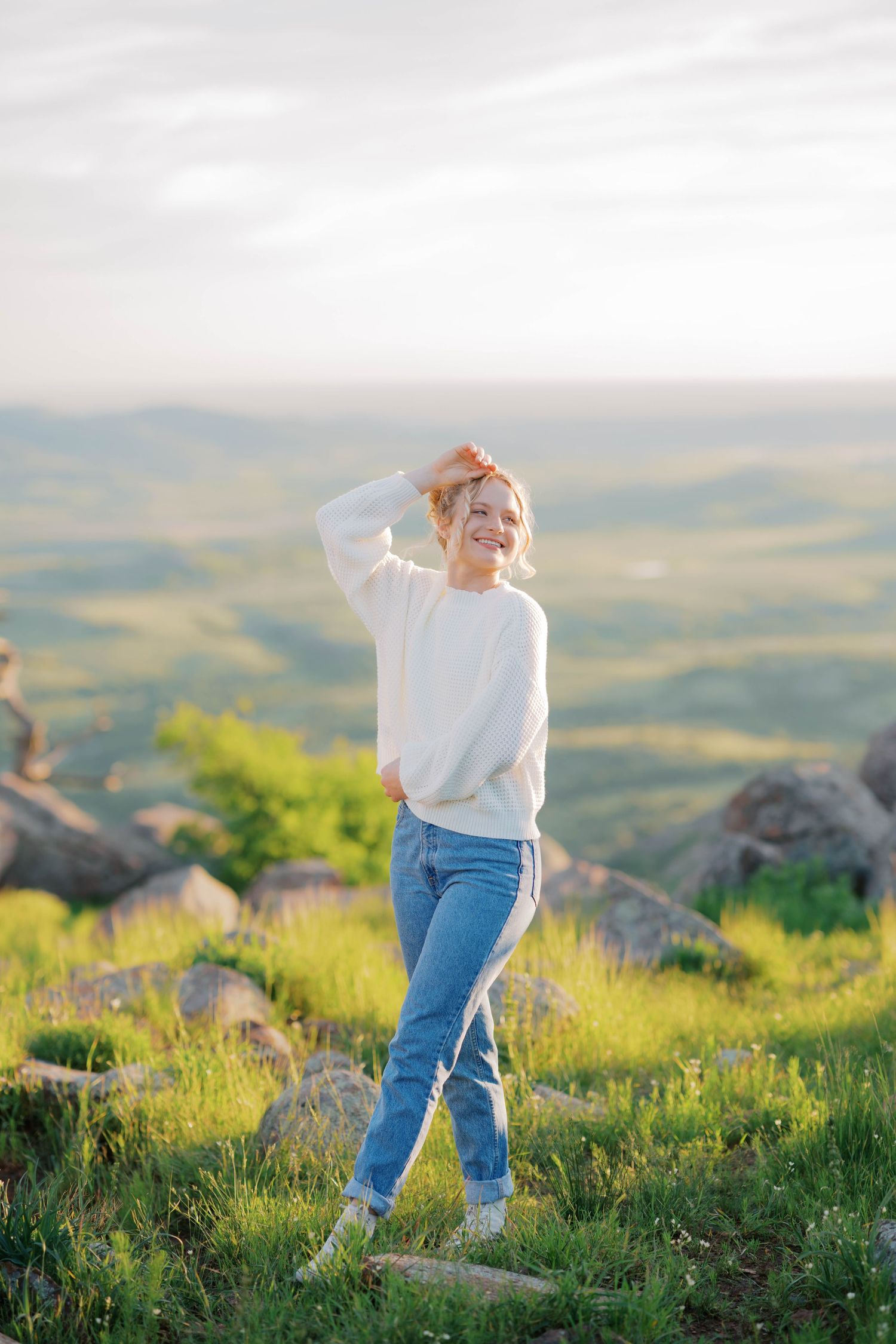 Smiling woman in white sweater and jeans poses on green hillside with scenic valley views in golden hour sunlight.