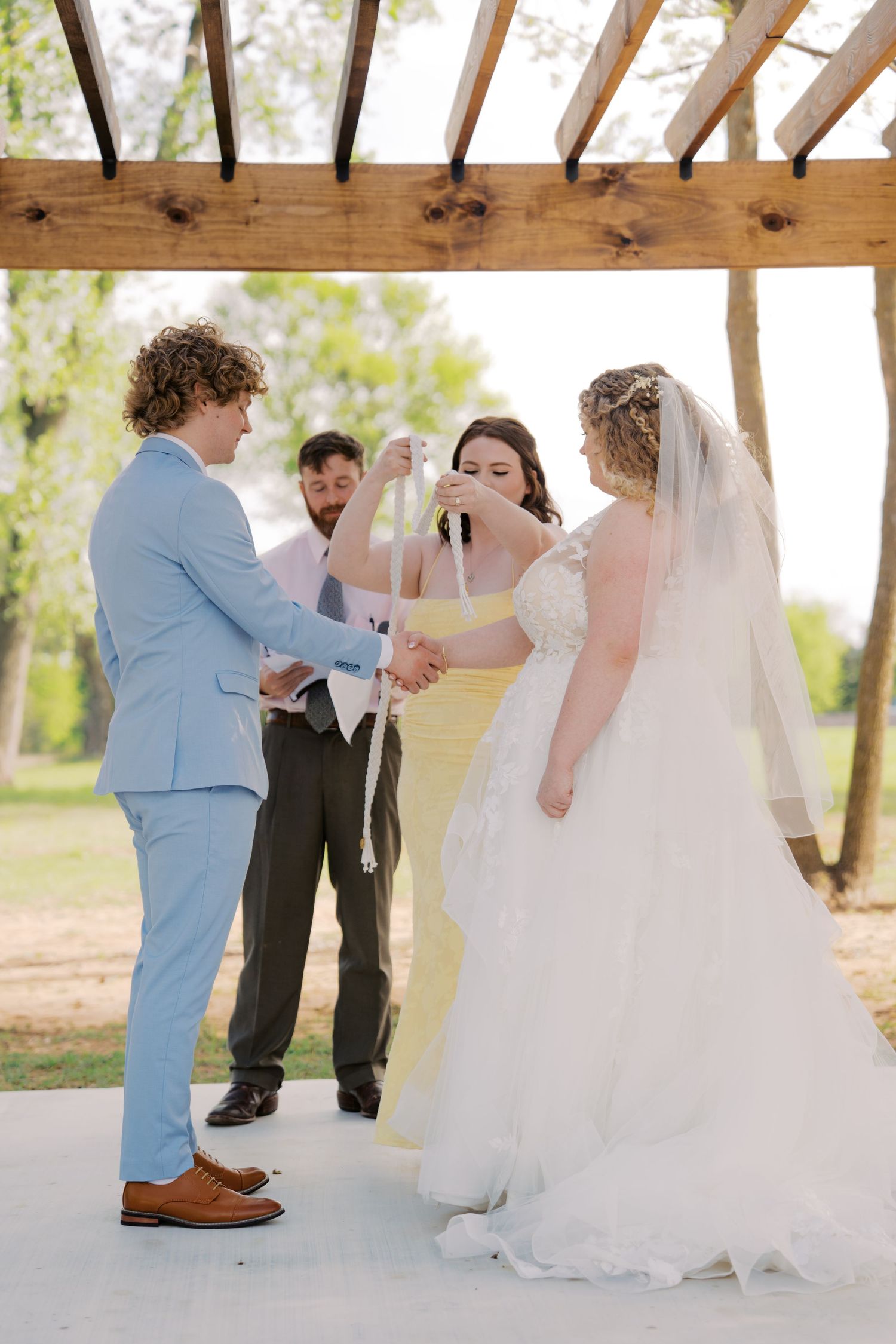 Bride and groom performing hand fasting ceremony under wooden pergola with officiant and wedding party.