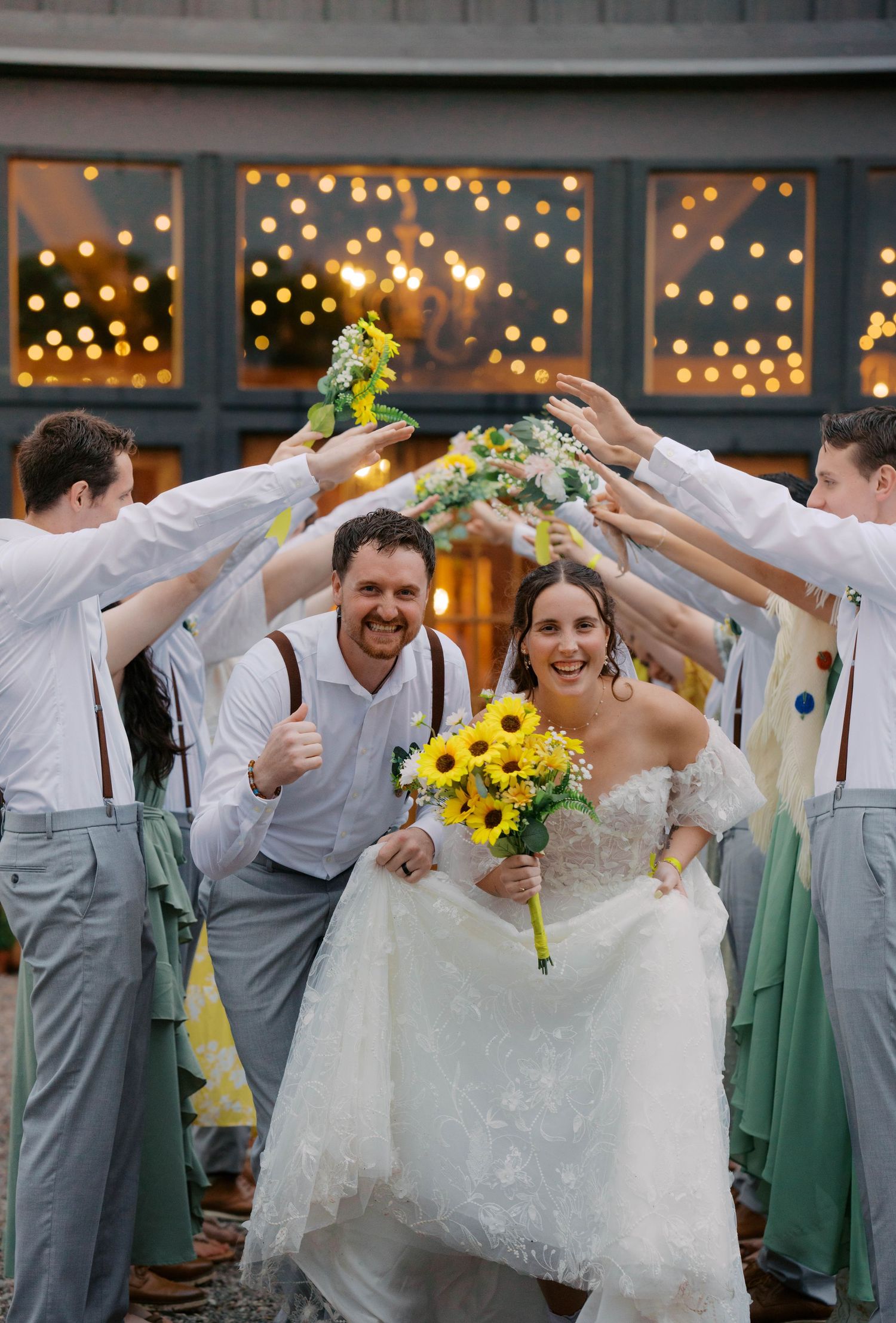 Smiling newlyweds walk through arch of raised arms formed by wedding party holding sunflower bouquets. Oklahoma wedding photographer.