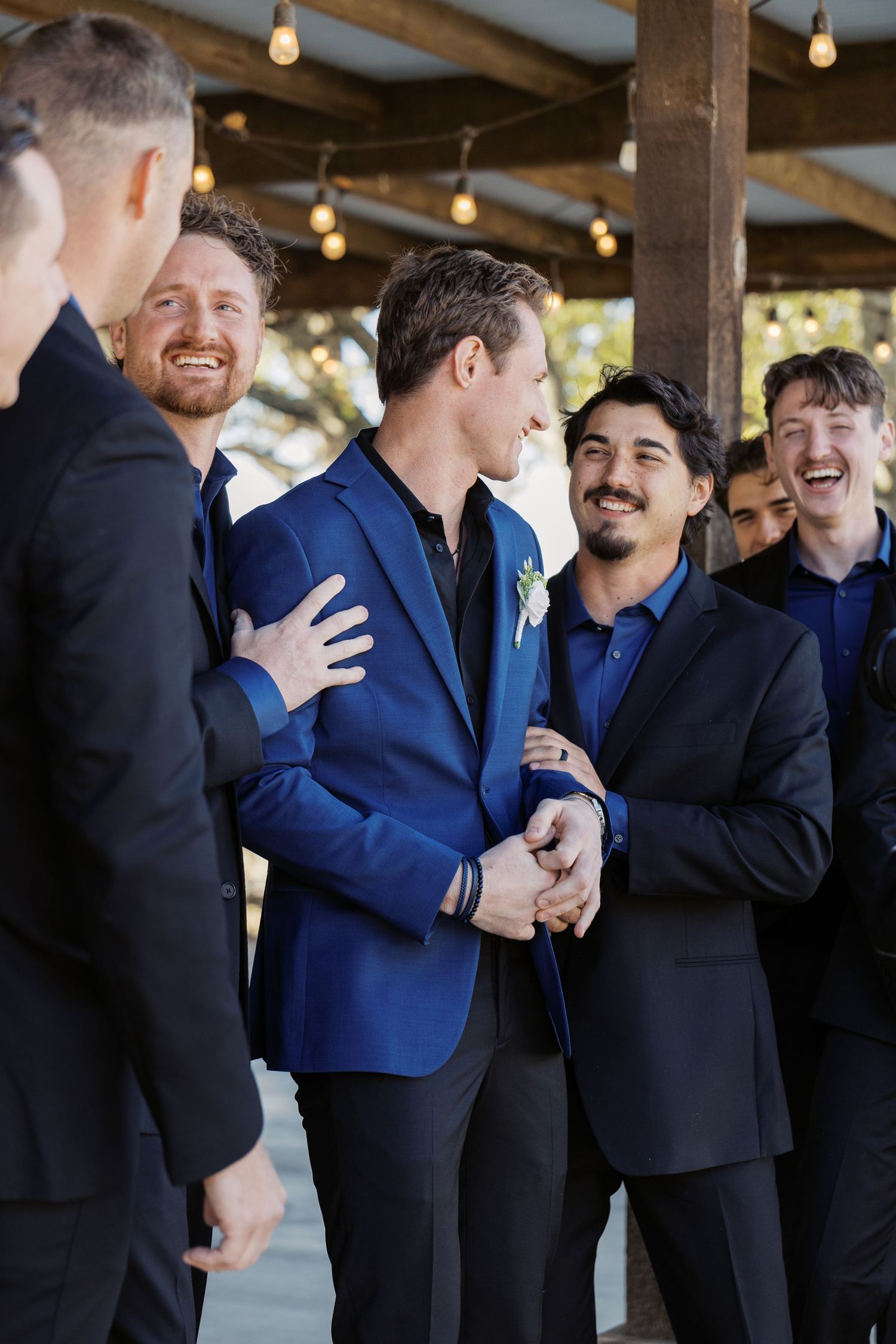 Groom in blue blazer laughing with groomsmen in black suits at outdoor wedding venue with string lights. Oklahoma wedding photographer.