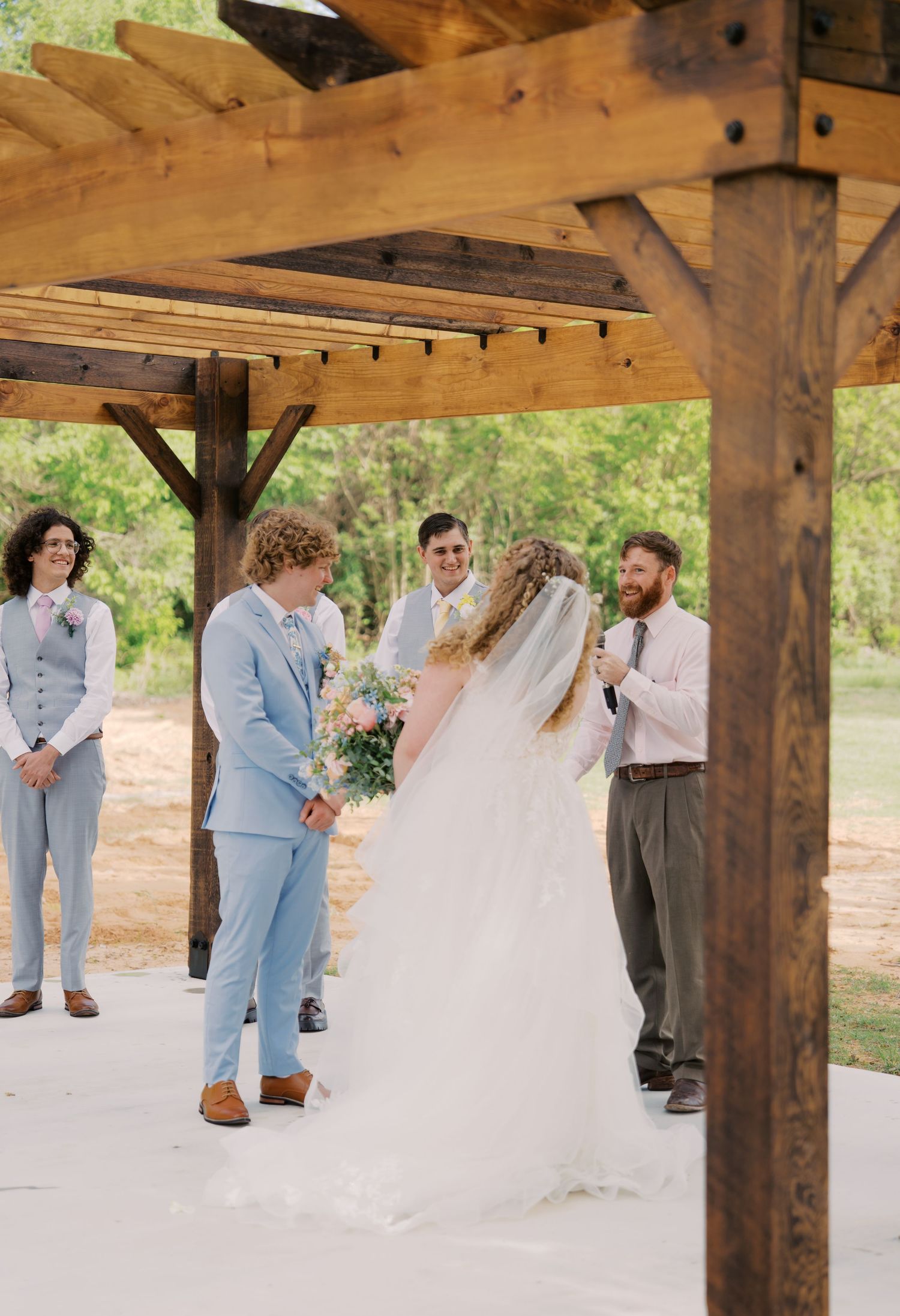 Outdoor wedding ceremony under rustic wooden pergola with bride in white gown and groom in light blue suit exchanging vows.