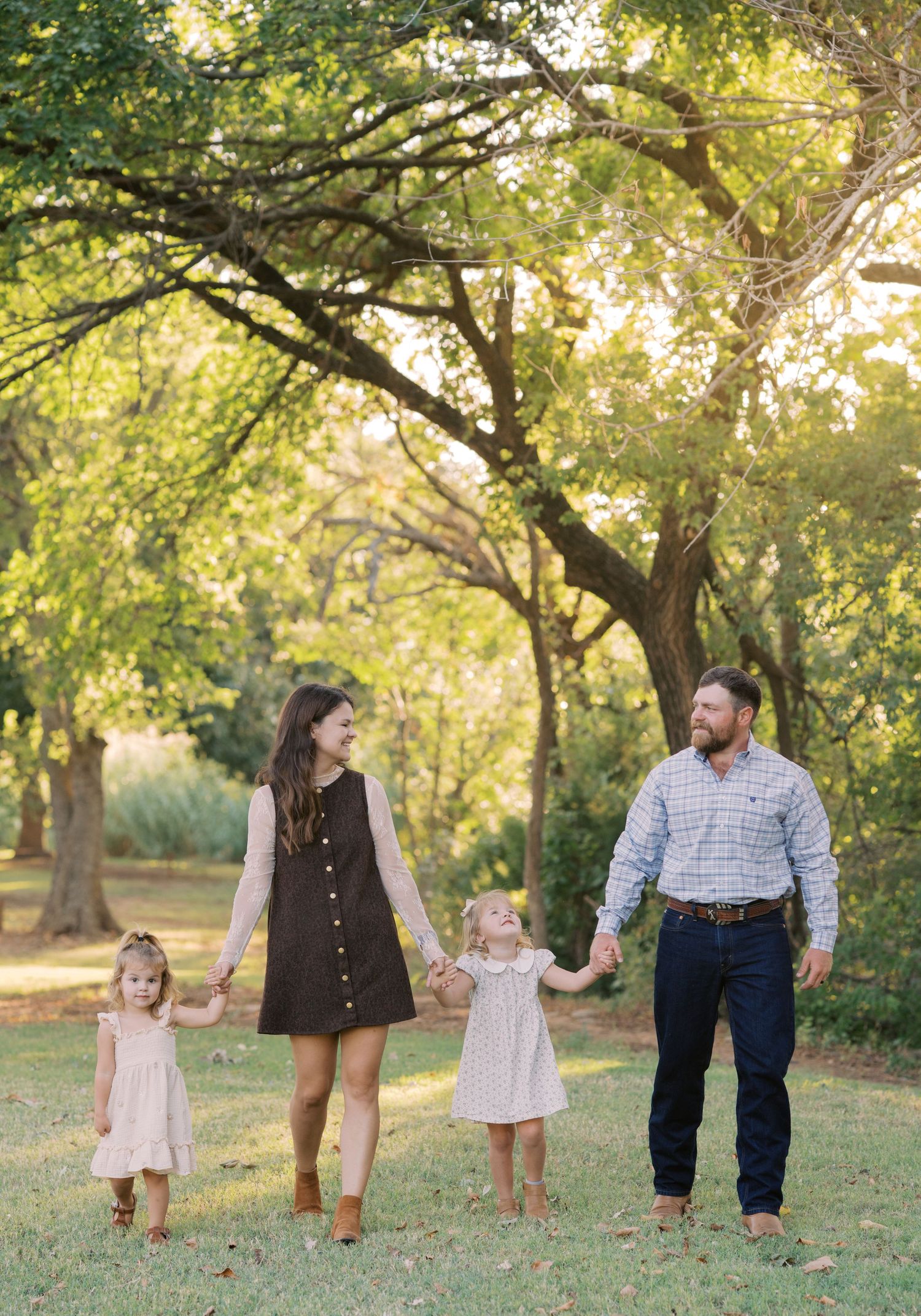 A family of four holds hands while walking through a sunlit park, with two young girls in white dresses and their smiling parents.