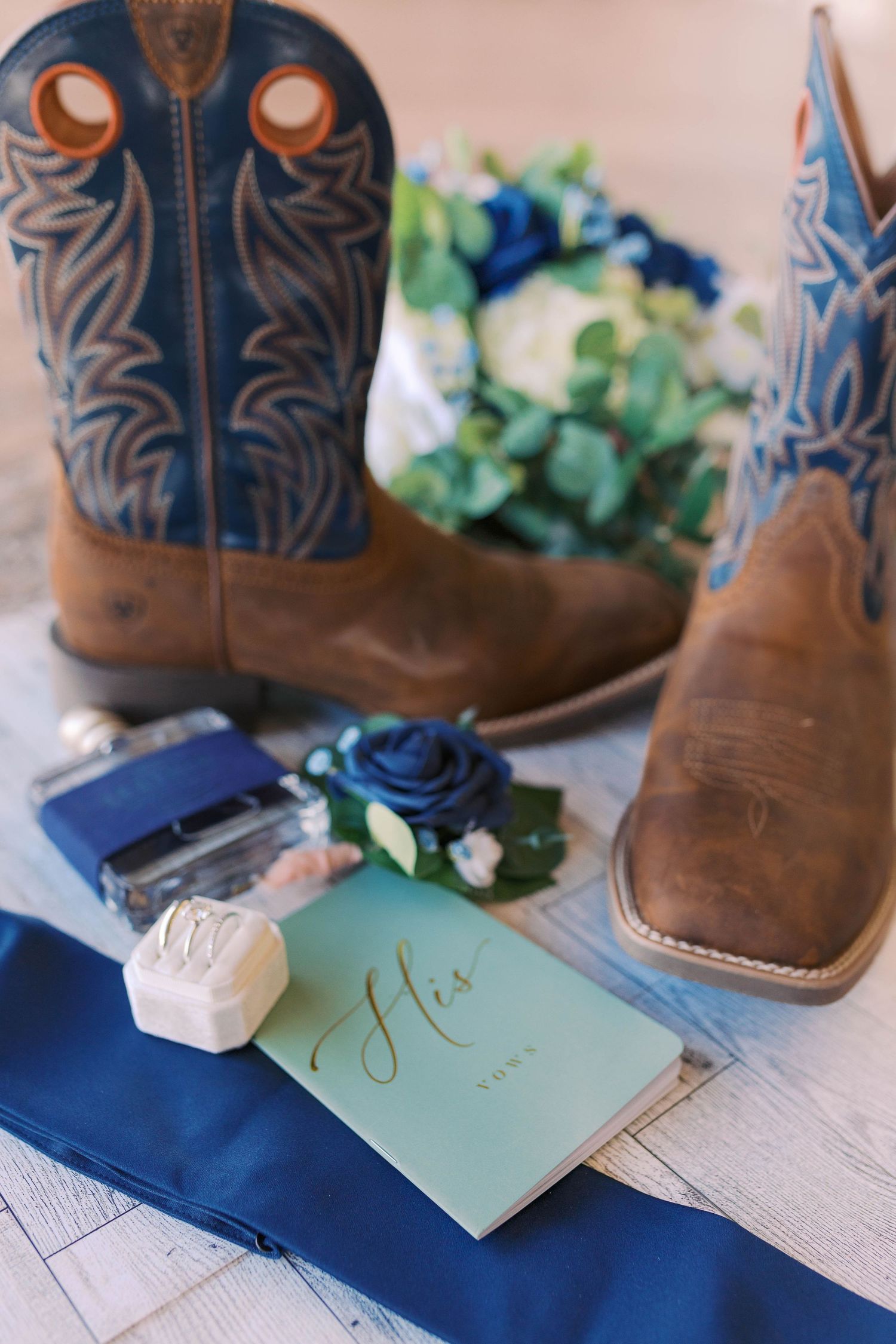 Groom wedding details including blue cowboy boots, boutonniere, vows booklet, ring box, and navy tie.