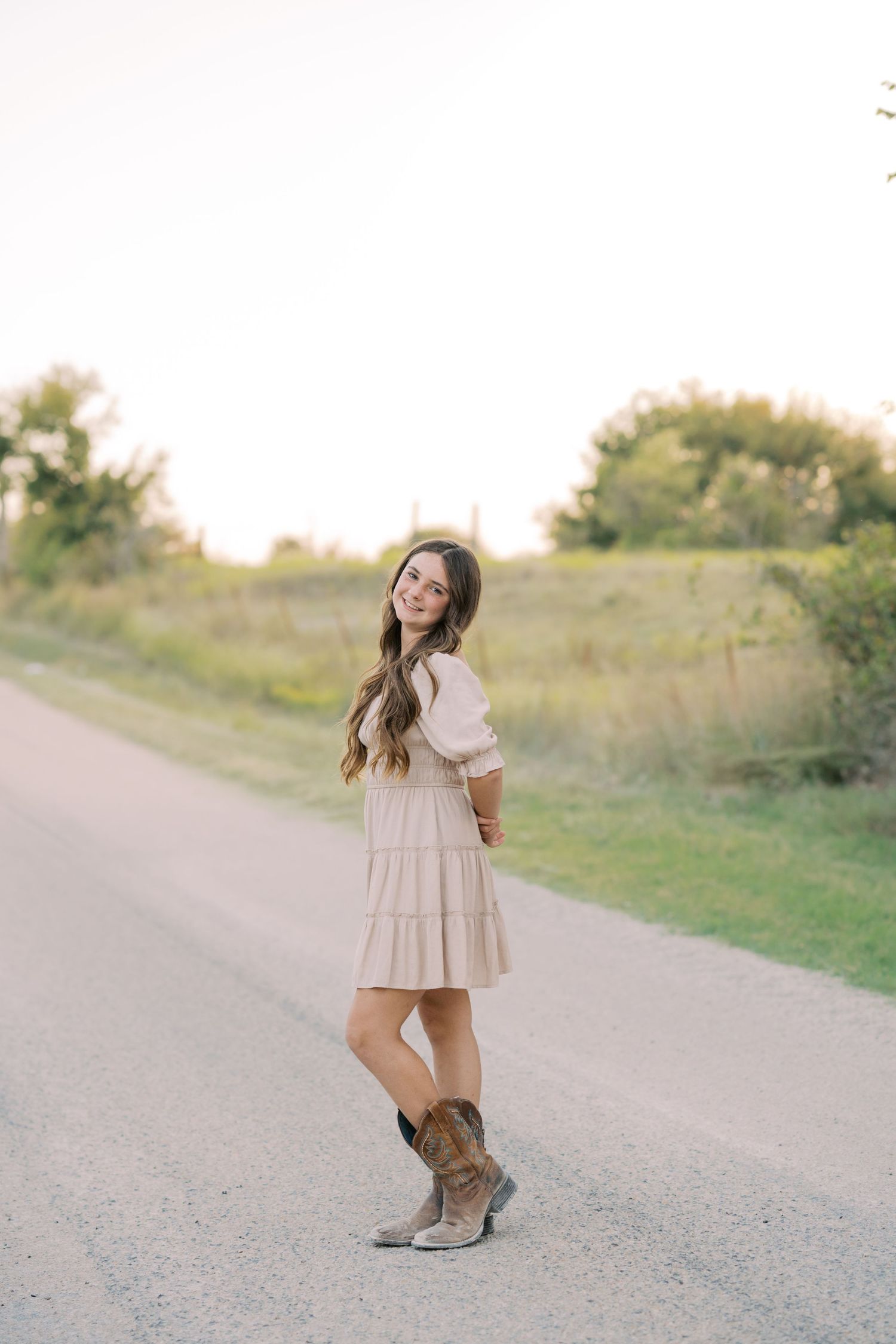 Young woman in beige tiered dress and cowboy boots smiling on a rural road surrounded by greenery.