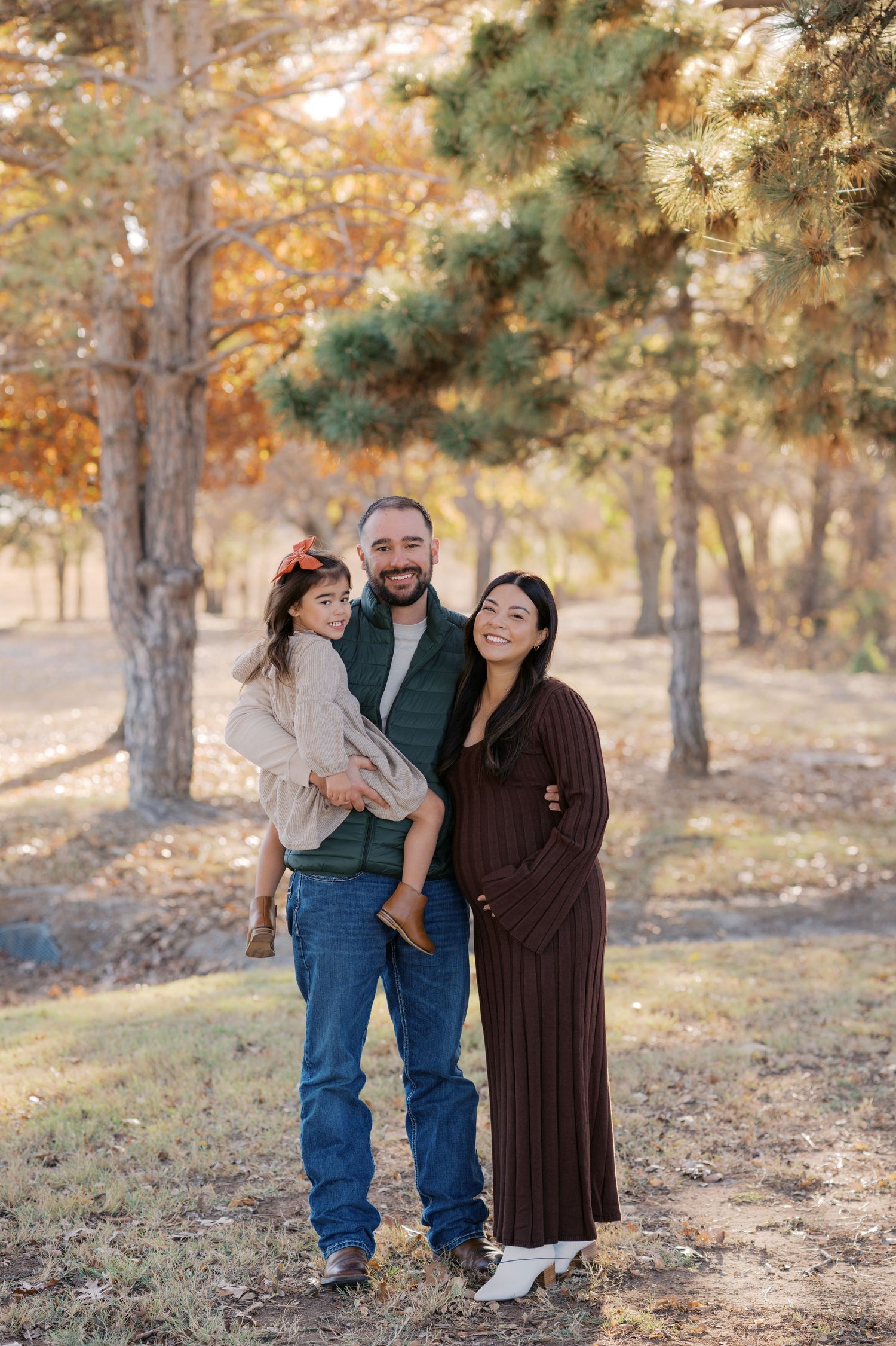 A happy family of three poses outdoors in autumn, man holding child, woman in brown dress, surrounded by colorful fall trees.