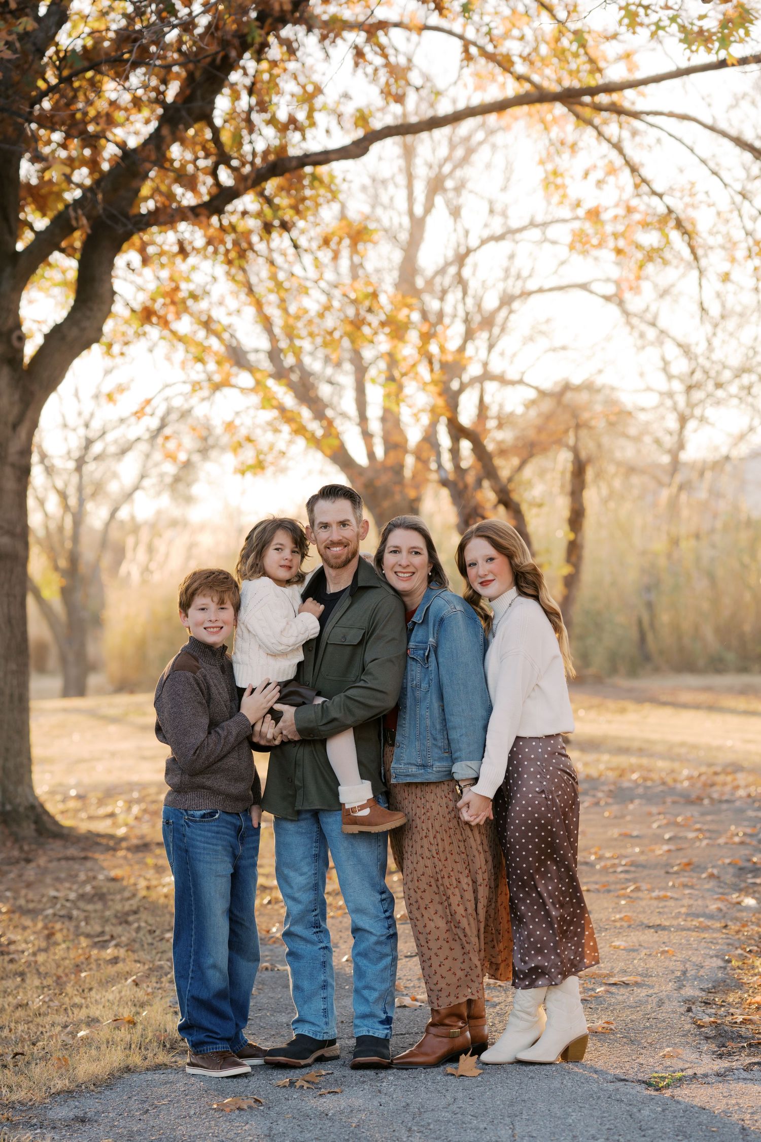 Family of five posing outdoors in autumn park with golden foliage, wearing casual fall outfits, smiling together on a path.