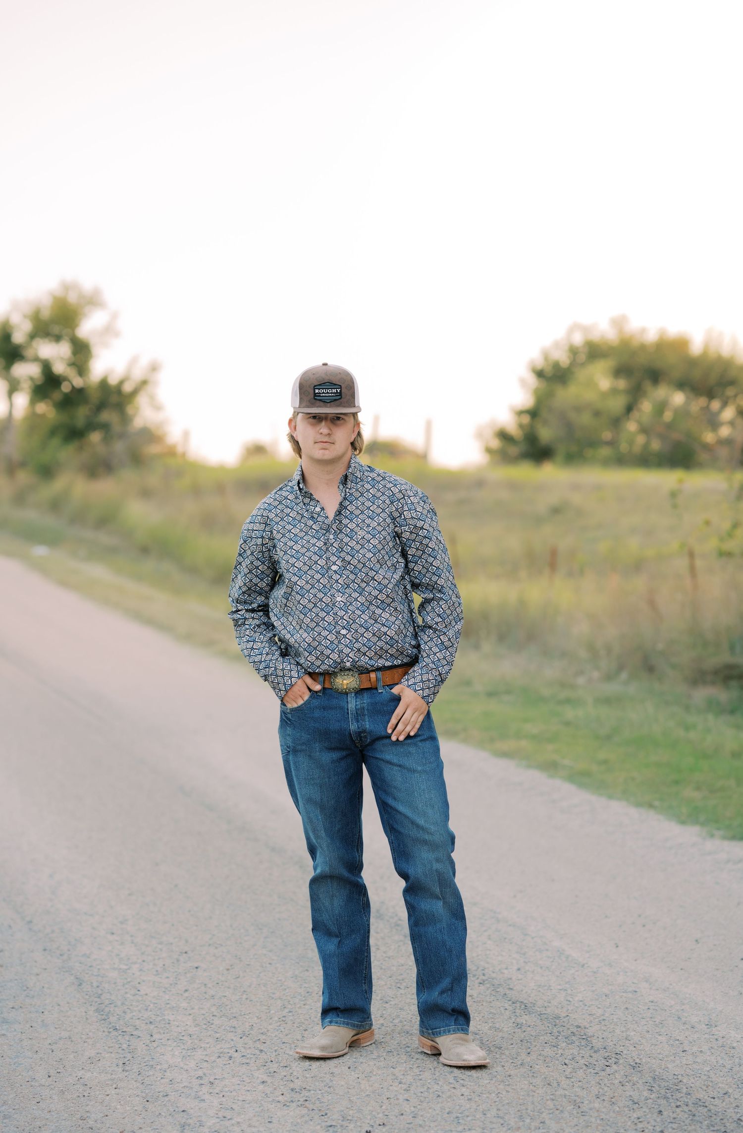 Young man in patterned shirt, jeans, and cap standing on a rural road surrounded by green fields.