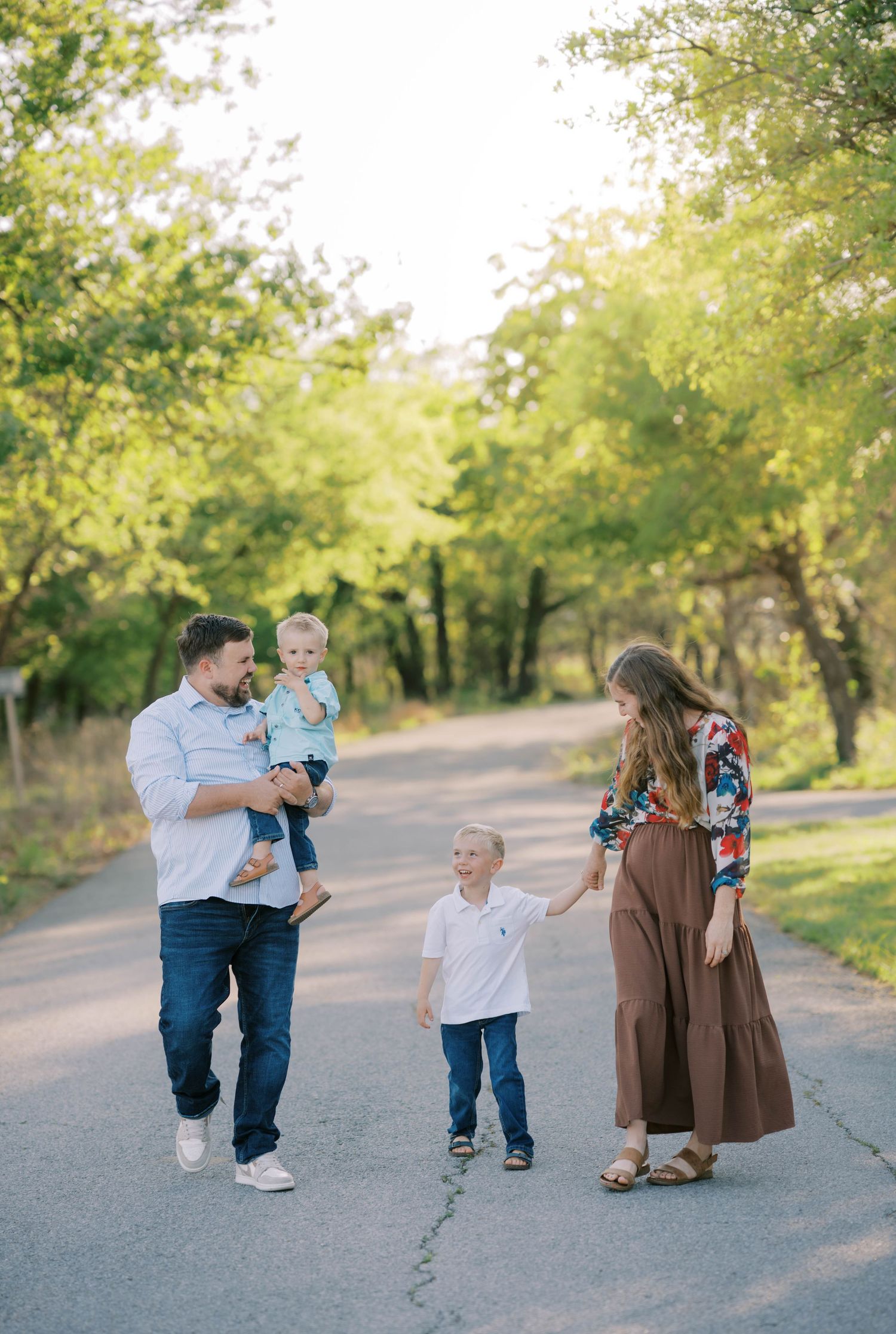Family of four enjoying a sunny walk on a tree-lined path, parents holding hands with young children outdoors.