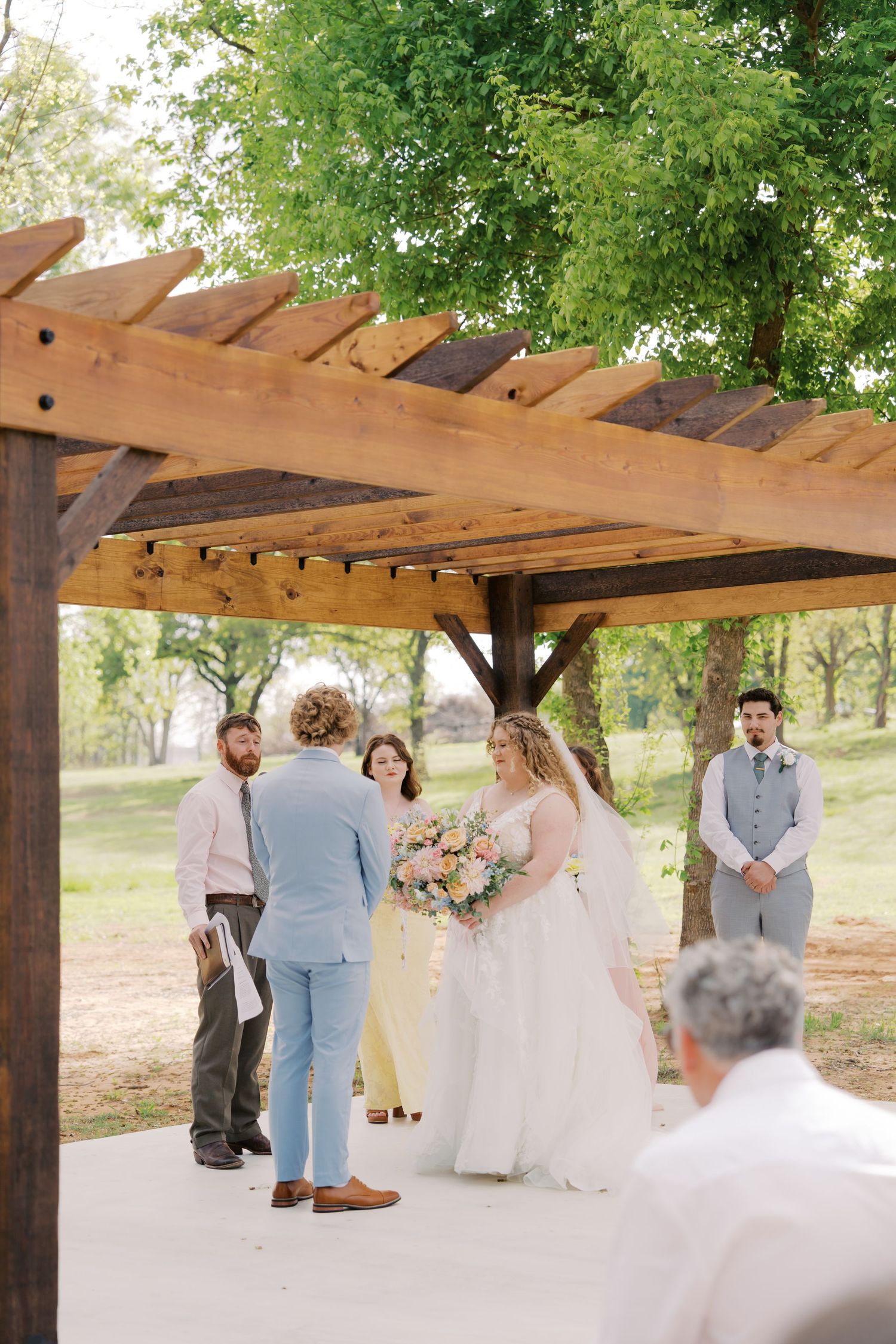 Bride and groom exchange vows under a rustic wooden pergola outdoors, surrounded by wedding party with lush green trees.