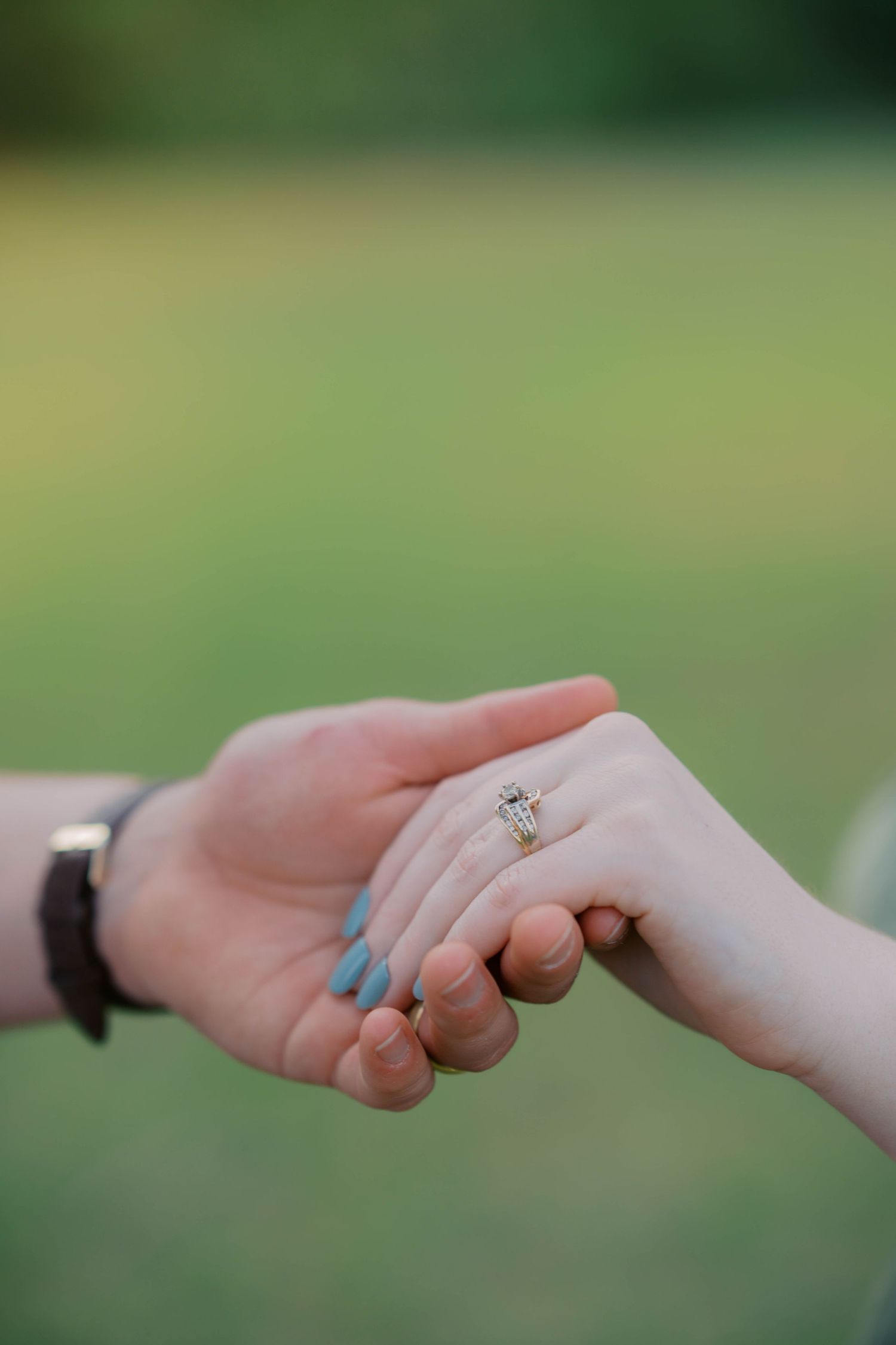 Close-up of two hands holding, woman wearing a diamond engagement ring with blue nail polish on green background.