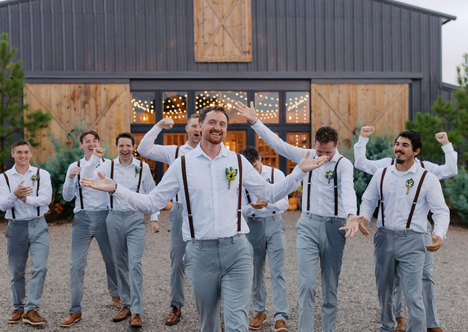 Groom and groomsmen in gray pants and suspenders celebrate outside a rustic barn wedding venue. Oklahoma wedding photographer.