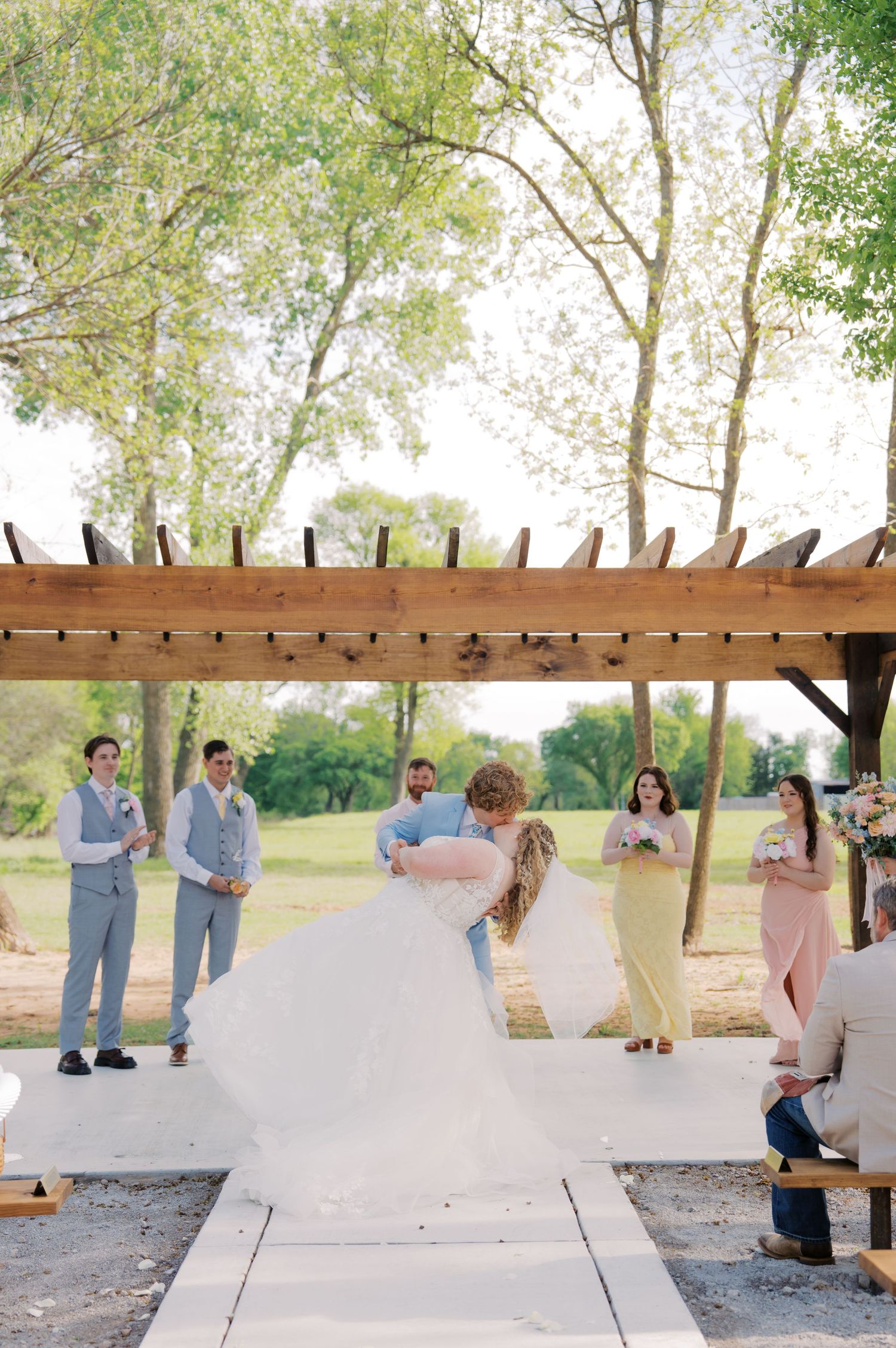 Bride and groom sharing first kiss under rustic wooden pergola during outdoor wedding ceremony.