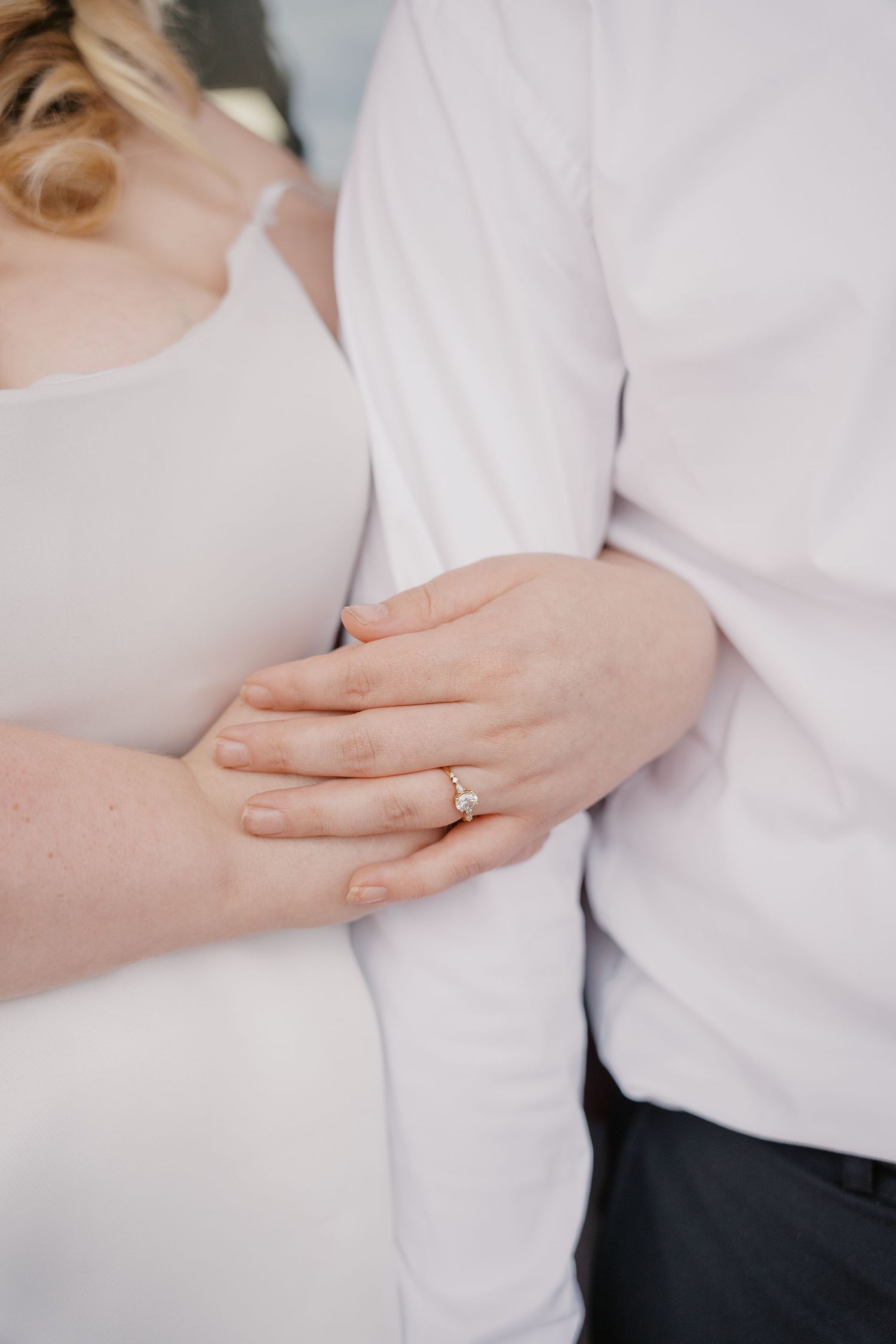 A bride wearing a diamond engagement ring gently holds her grooms arm in white attire.