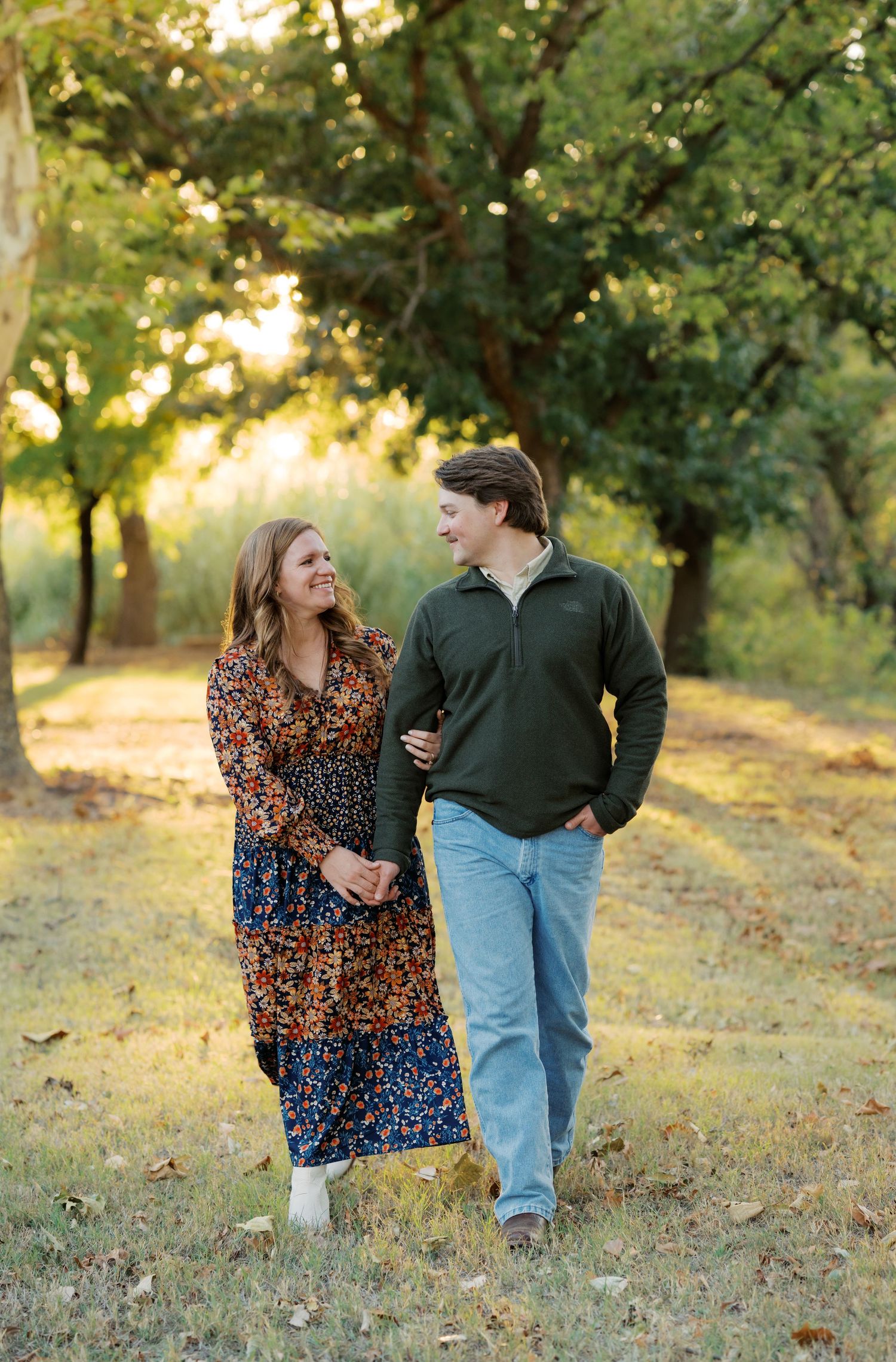 Laughing couple walking hand in hand through a sunlit autumn park during engagement photo session.