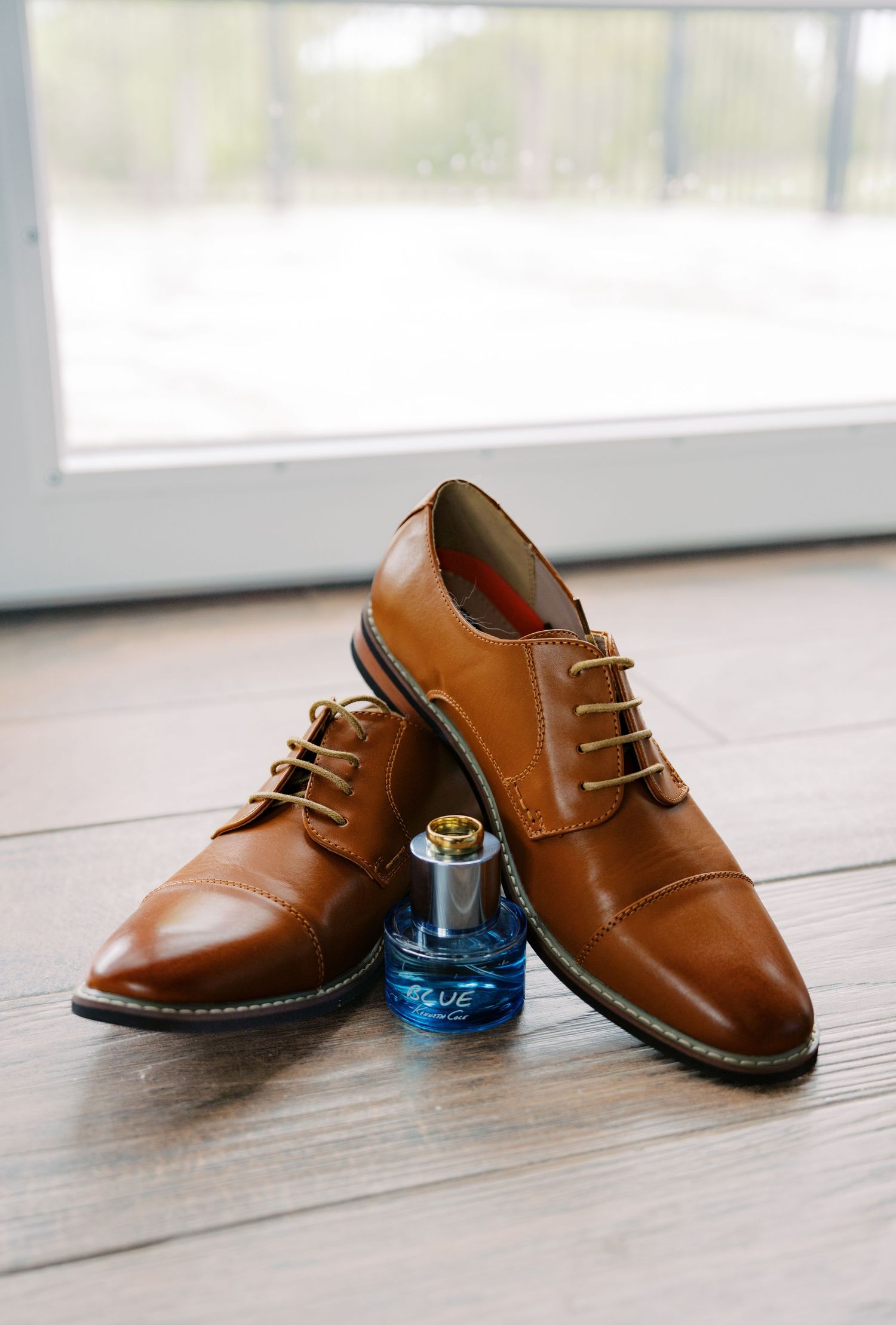 Brown leather cap-toe oxford dress shoes with tan laces beside a blue cologne bottle on hardwood floor.