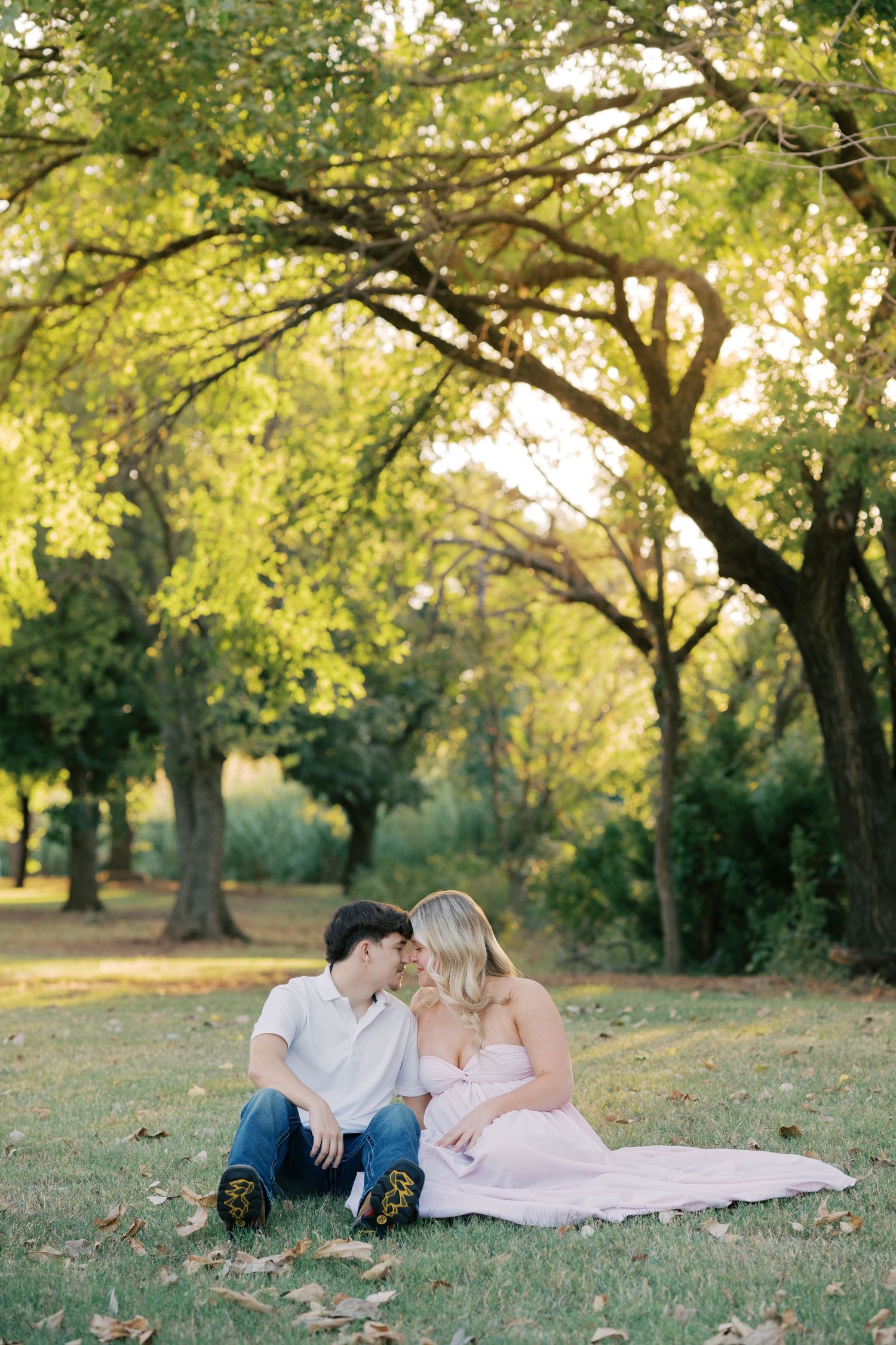 Expecting couple sits together on grass under sunlit trees in a tender maternity photo session.