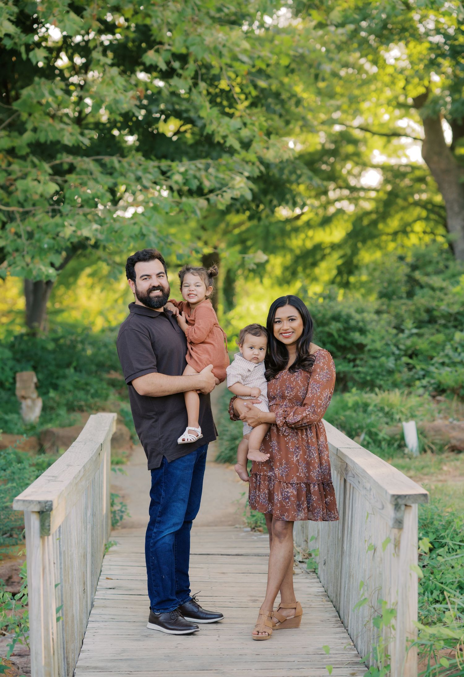 A family of four poses on a wooden bridge surrounded by lush green trees, with parents holding two young children.