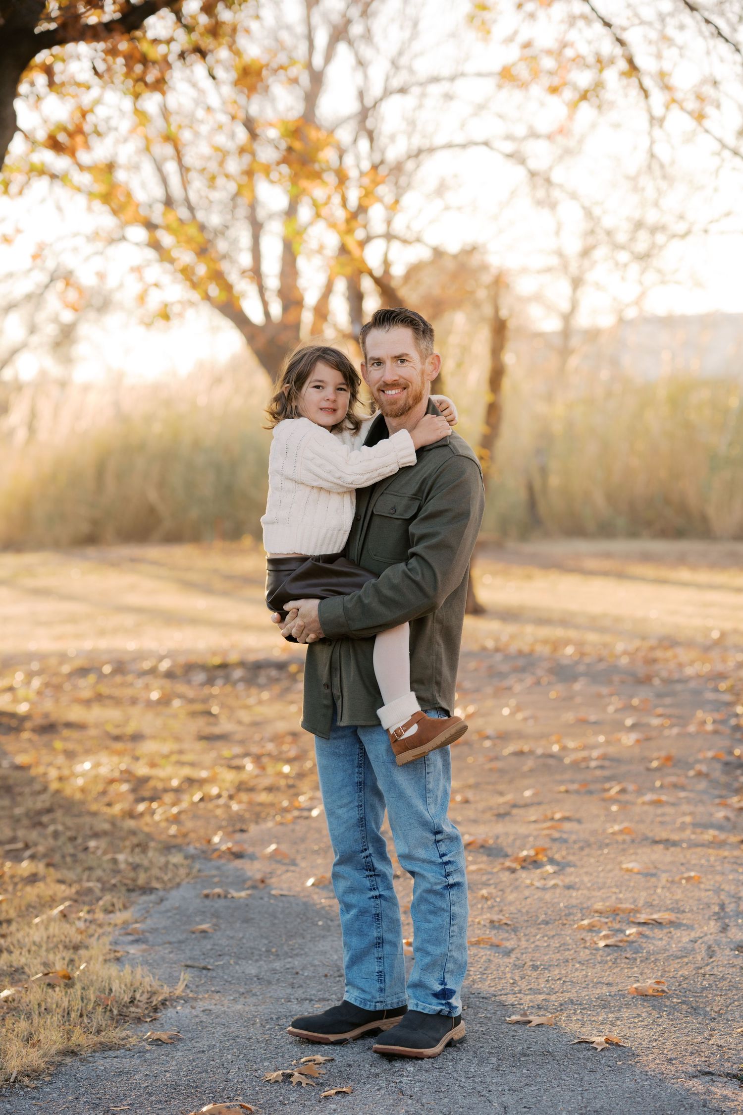 A father holds his young daughter in his arms outdoors during golden hour in autumn, both smiling warmly.