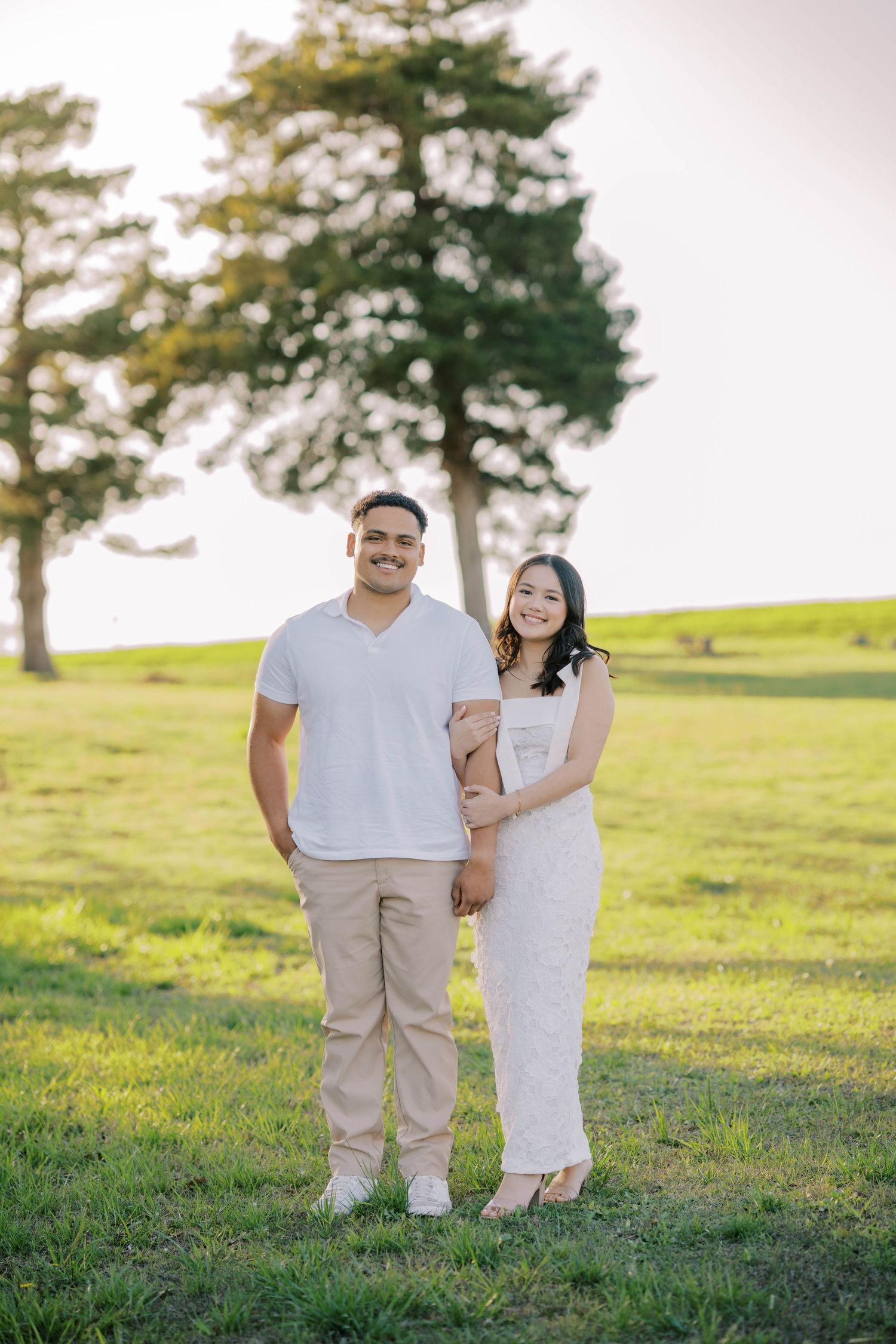 Smiling couple poses together on a green lawn with trees in the background during golden hour.