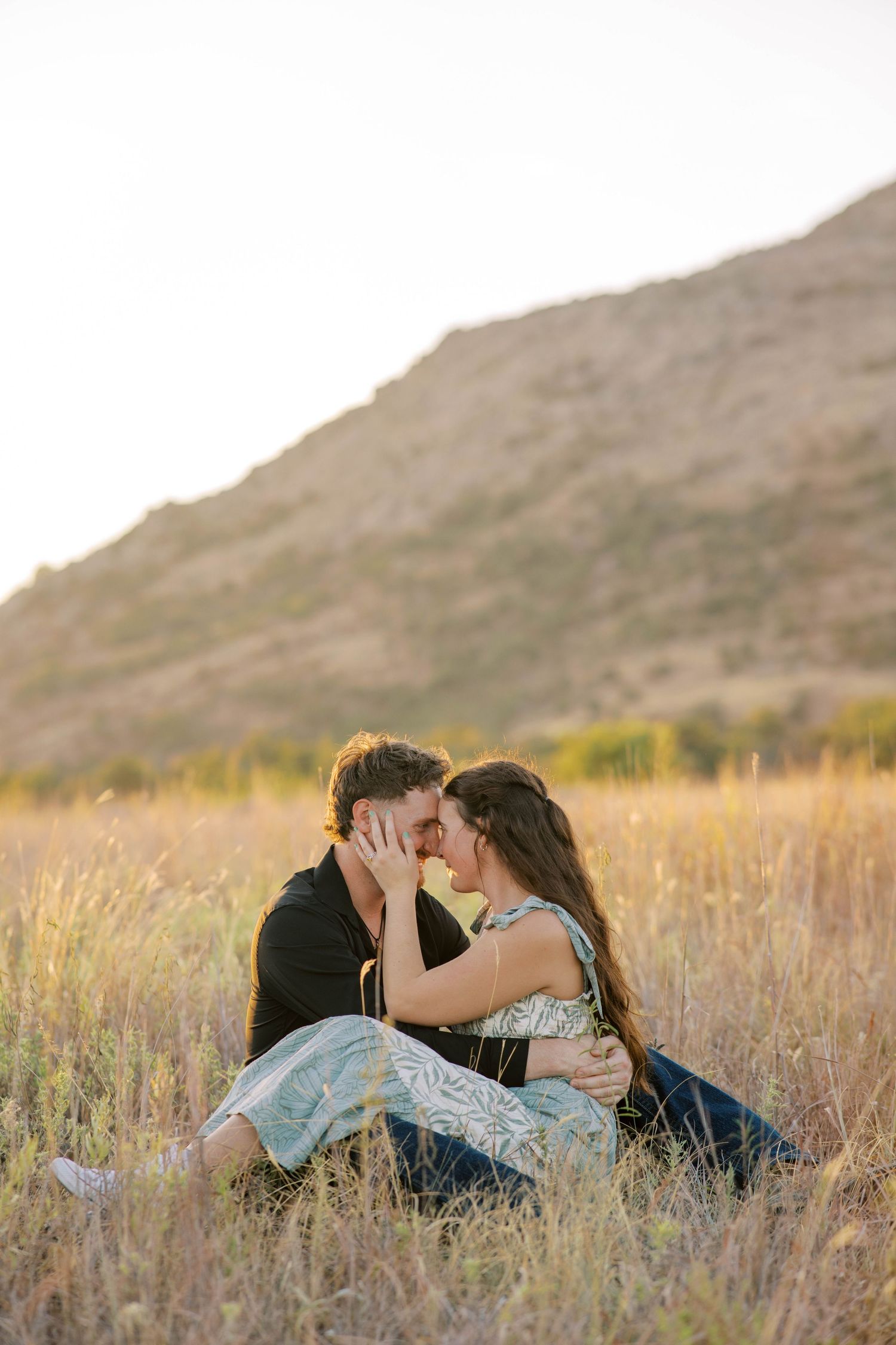 Couple sharing a romantic moment sitting in golden grass field with a scenic mountain backdrop at sunset. Oklahoma wedding photographer.