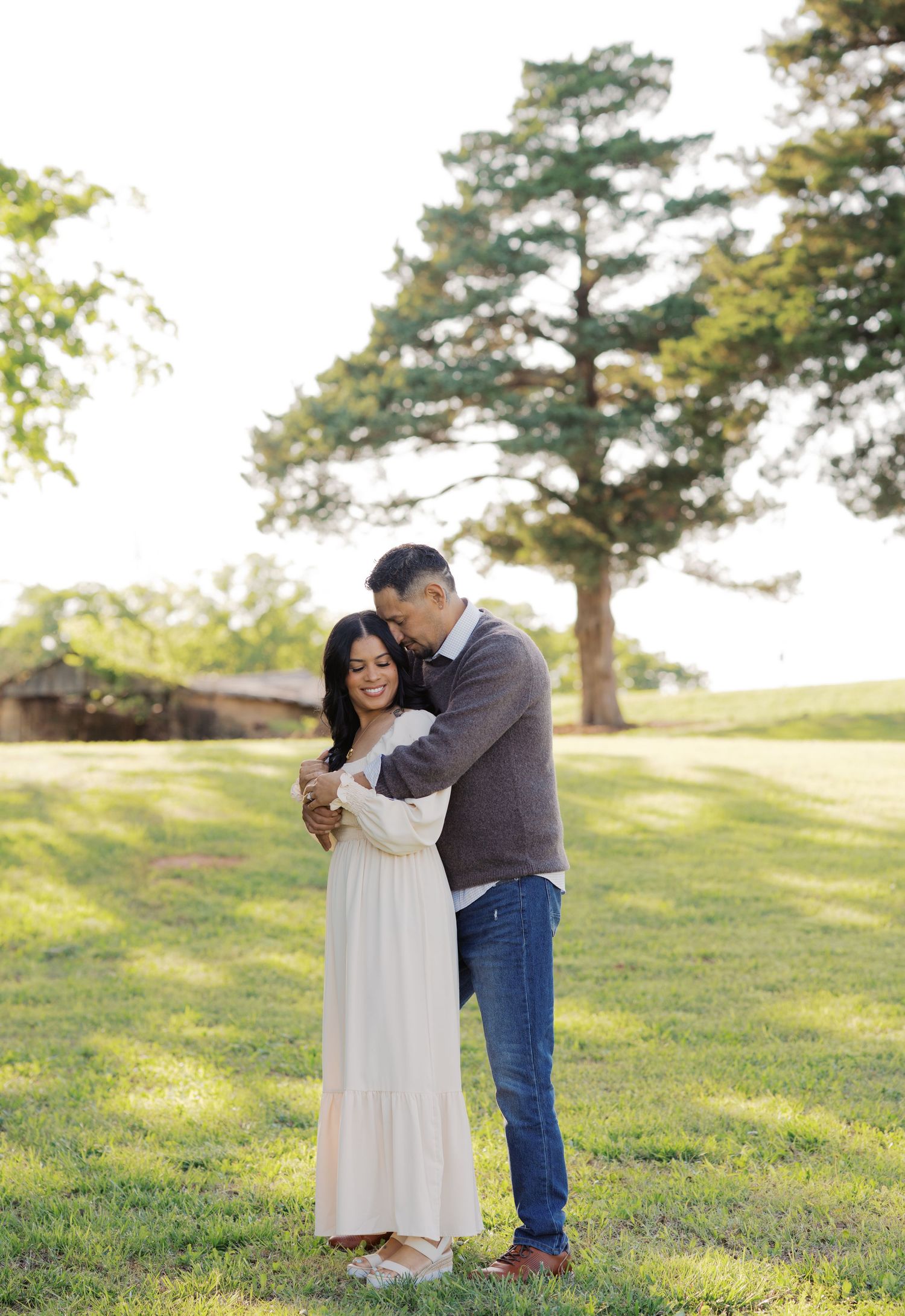 A couple embraces on a green lawn, the man hugging the woman in a white dress from behind, smiling near a large tree.