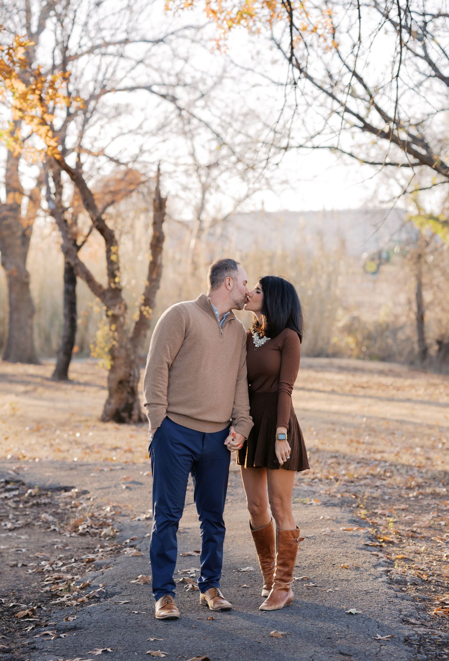 Couple sharing a kiss while holding hands on a scenic autumn path surrounded by golden trees.