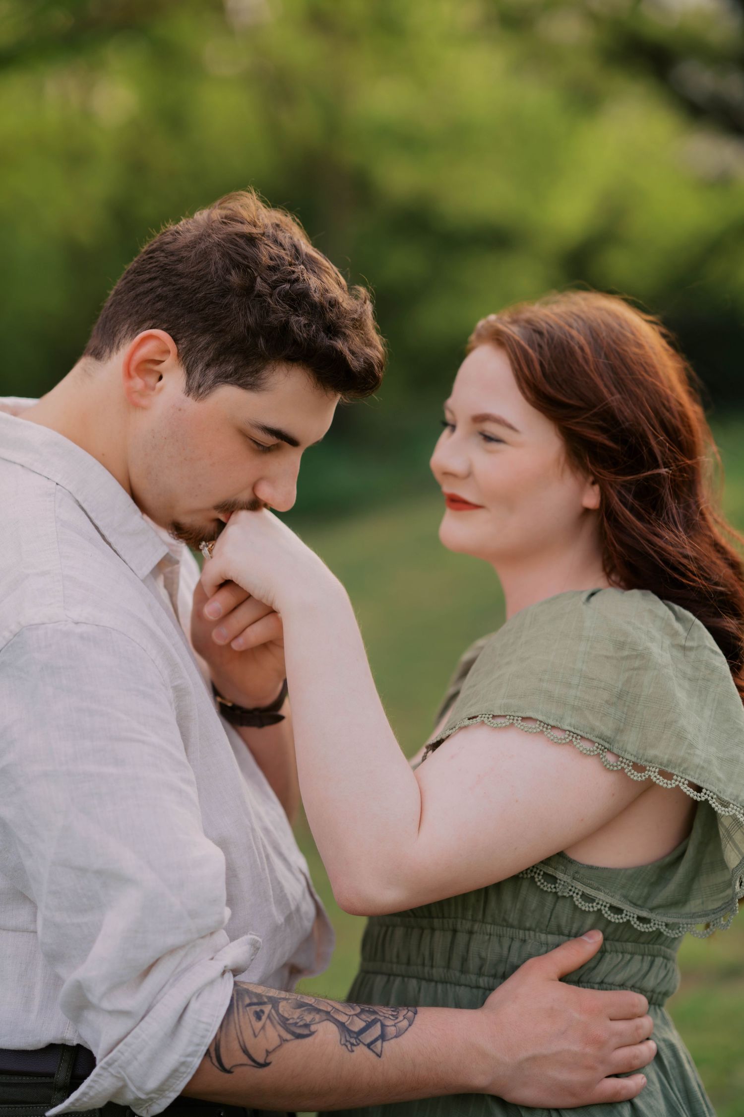 A man kisses a woman's hand outdoors, as she smiles warmly, both dressed in soft green and white tones.