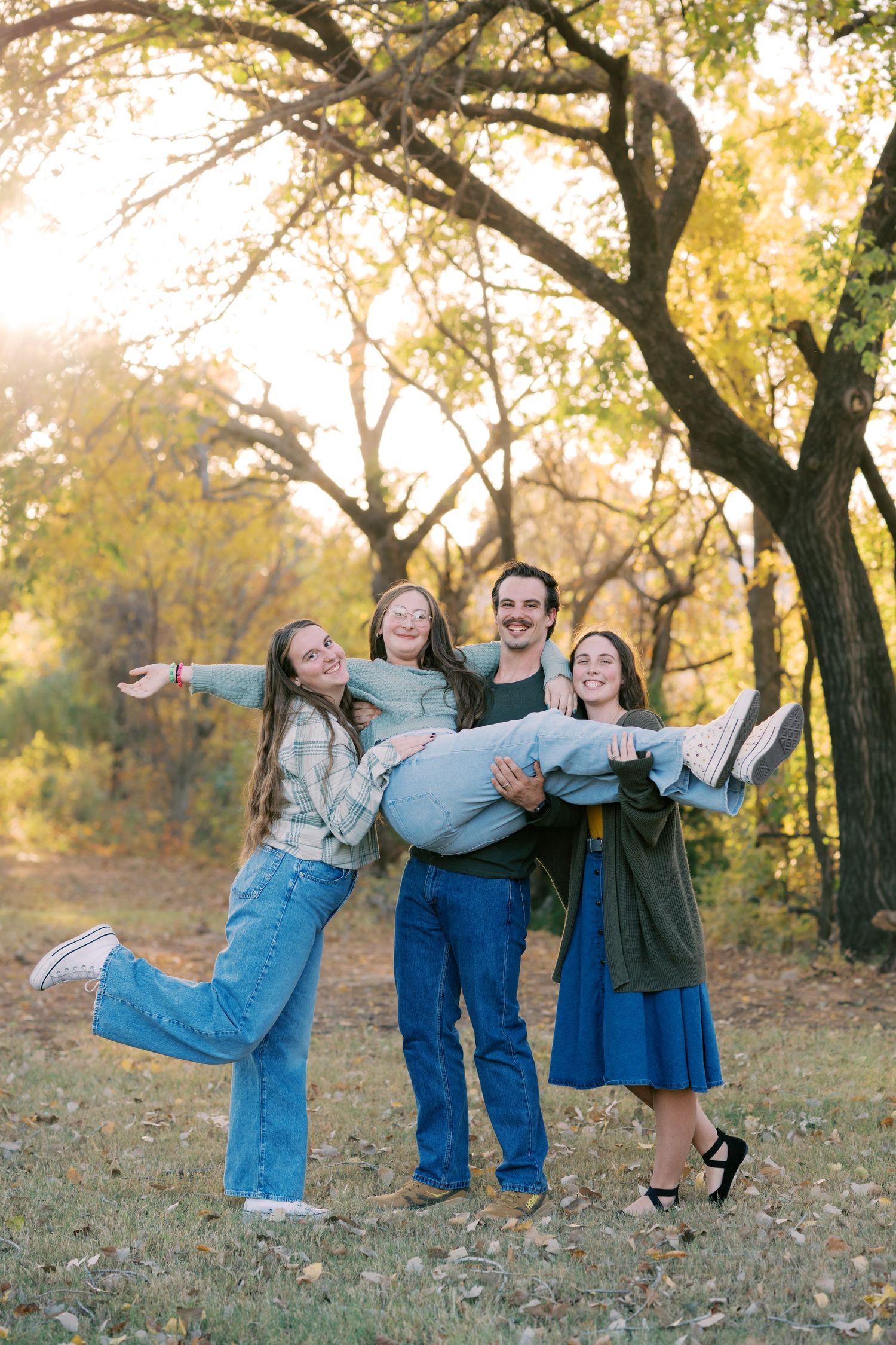 Joyful family posing playfully in autumn park, man lifting woman while two others laugh beside them.