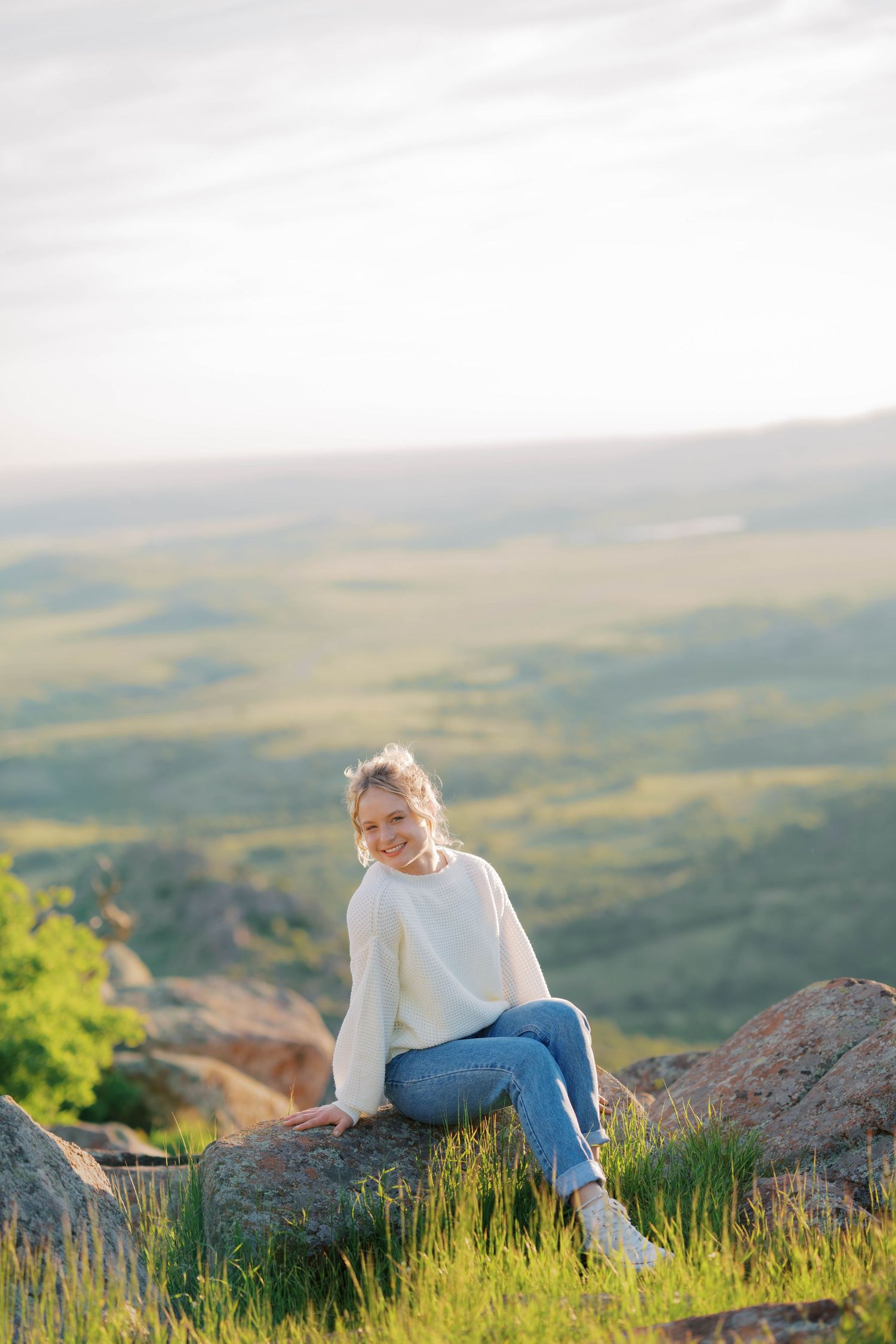 Young woman in white sweater and jeans sitting on rocks with scenic green valley landscape in background.