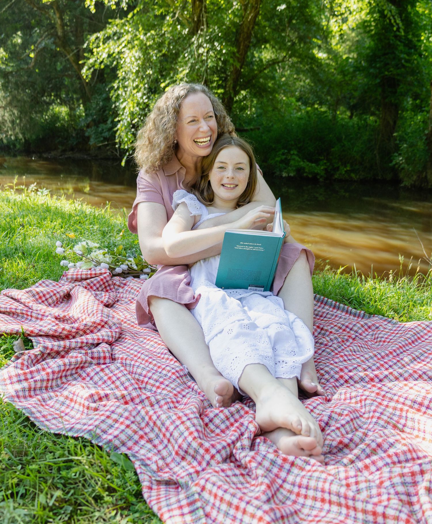 A group enjoys a sunny outdoor picnic on a red and white checkered blanket near a stream in a lush green park.