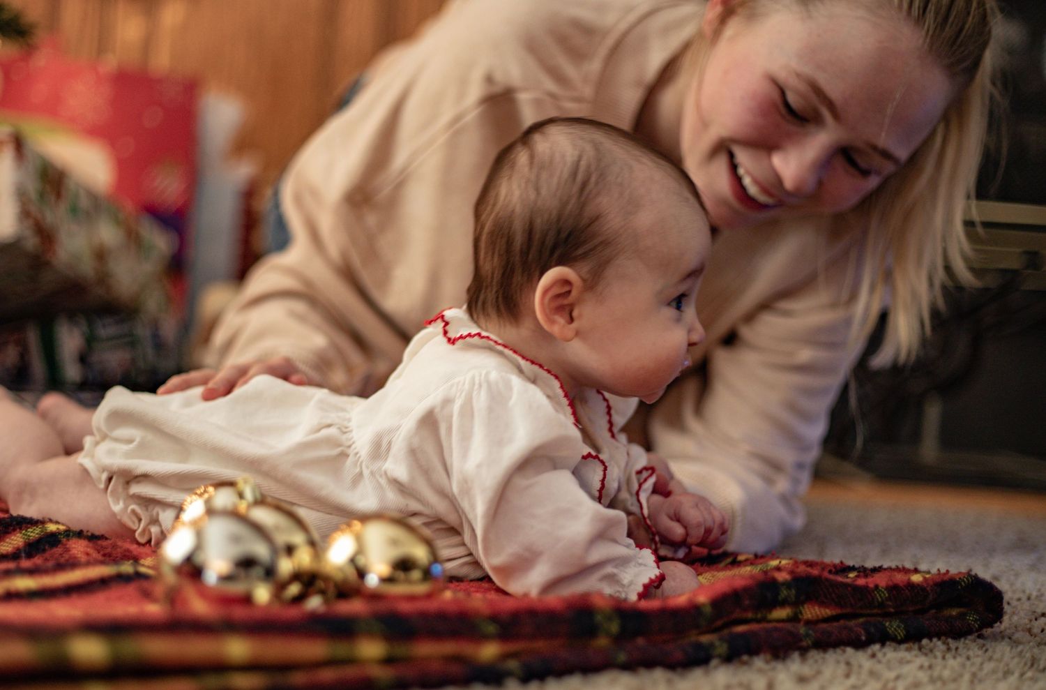 A baby in a white outfit lies on a patterned rug while someone smiles warmly nearby.