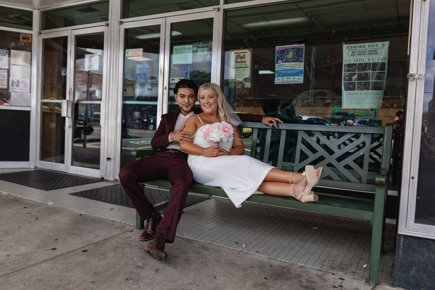 A couple sits together on a green bench outside a storefront.