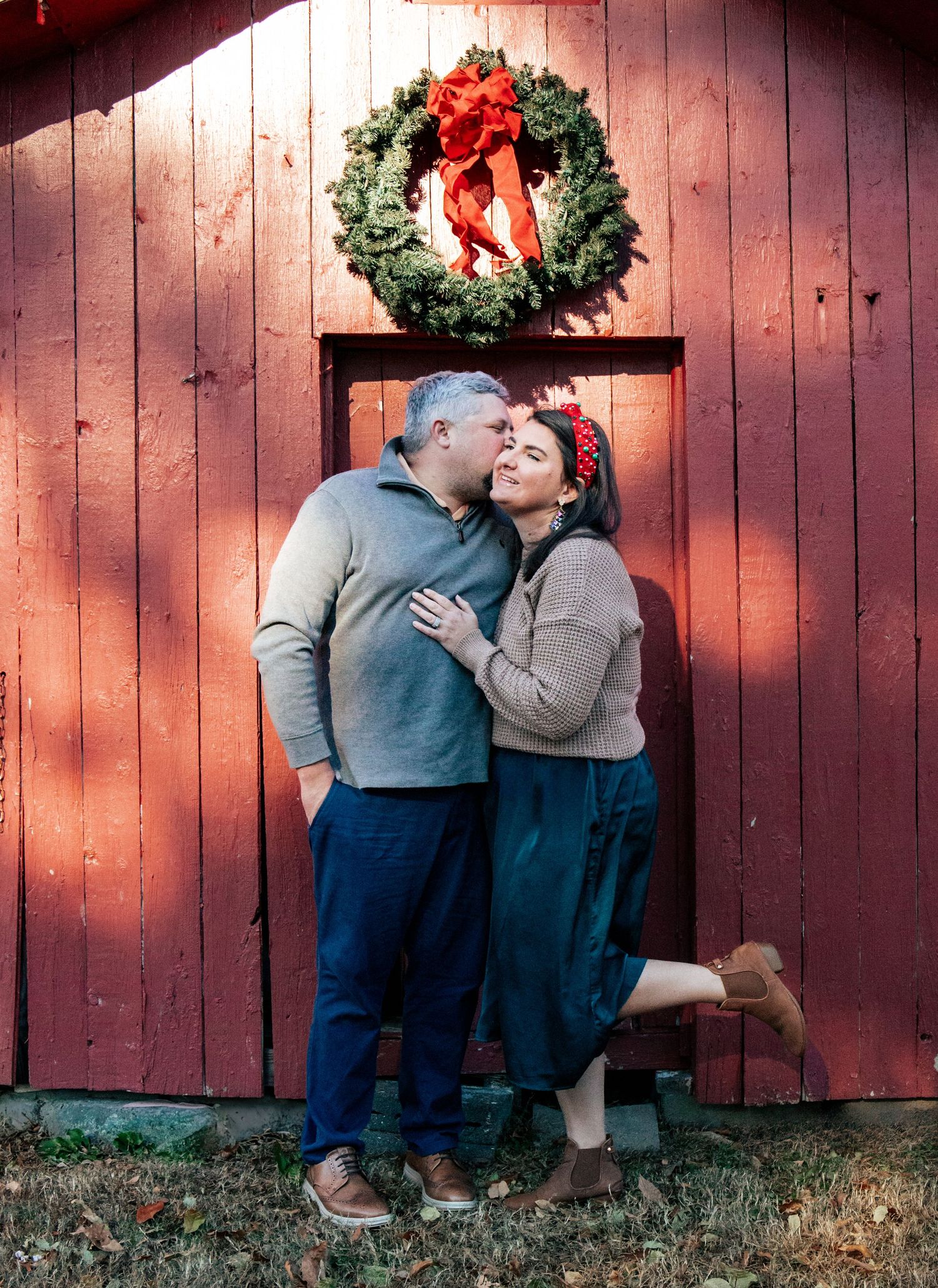 Couple poses affectionately against red barn wall with holiday wreath decoration.