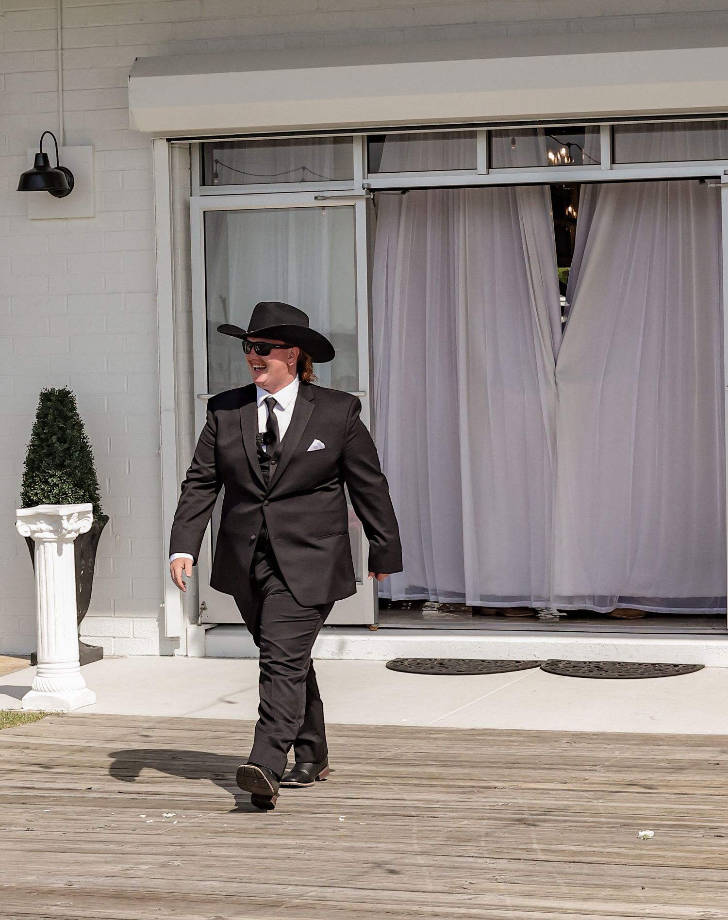 Groomsmen in black suits and cowboy hats walking on a wooden boardwalk near a white building.