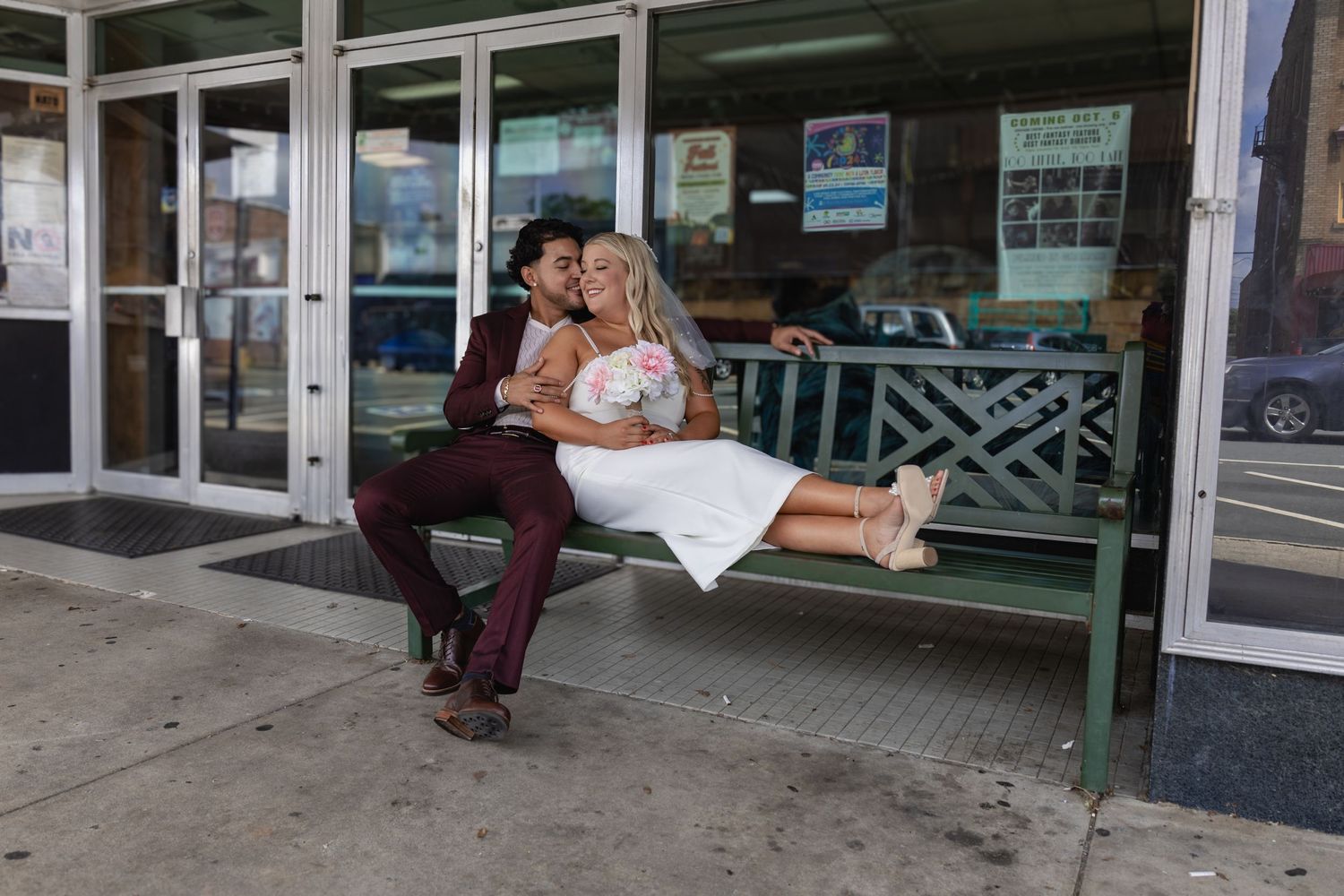 A couple shares a romantic moment on a green bench outside a shop.