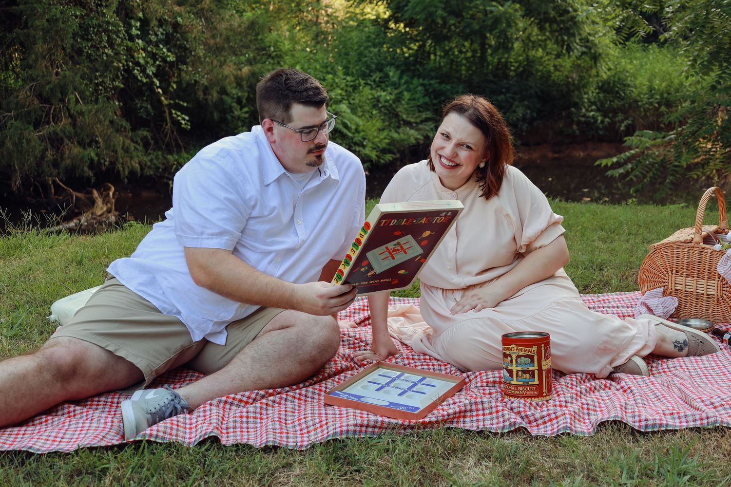 A couple enjoys a romantic picnic on a red checkered blanket with books and snacks in a garden setting.