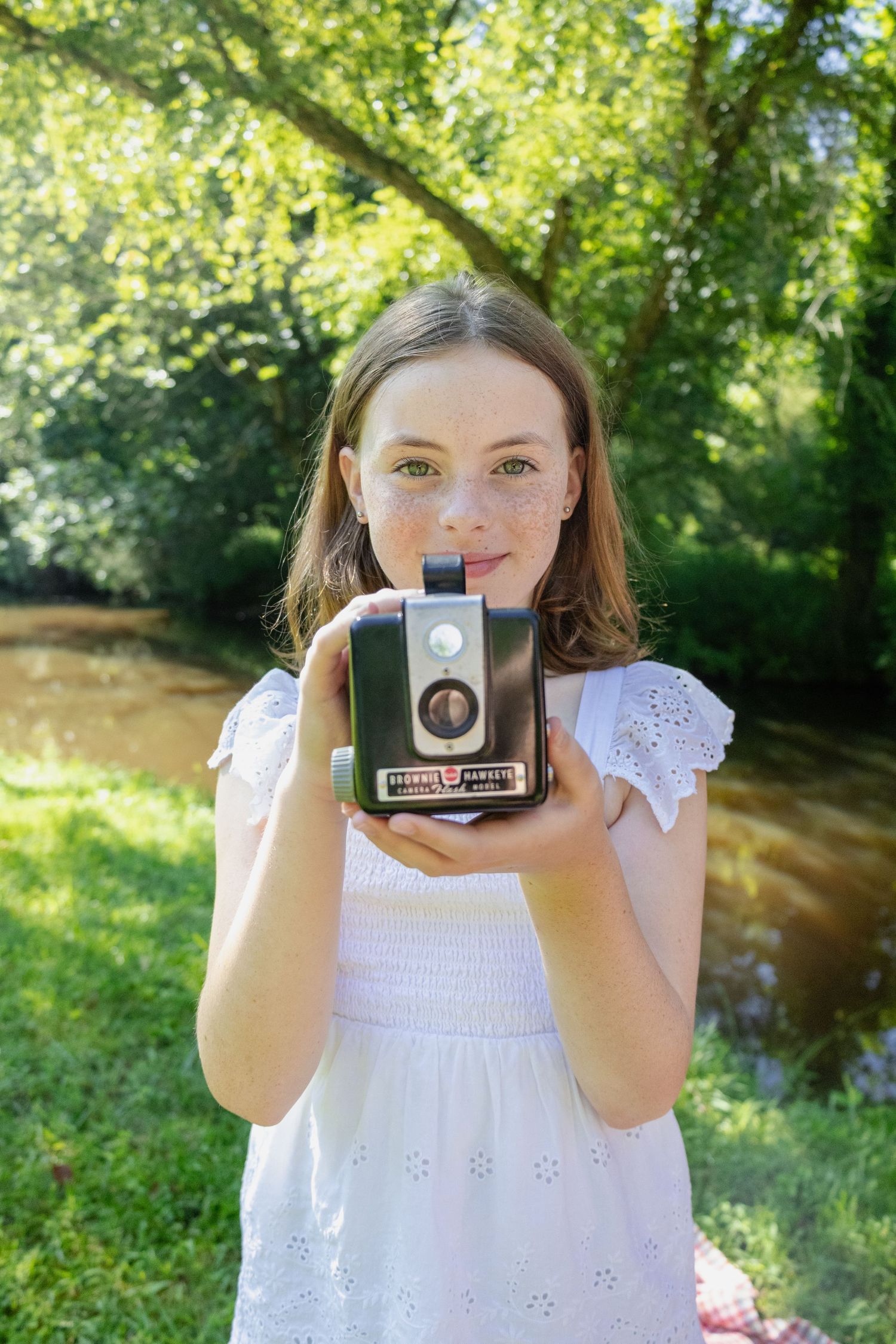 A vintage camera is held up in a sunny outdoor setting with greenery in the background.