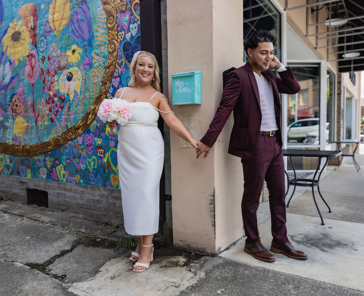 A couple in wedding attire holds hands in front of a colorful floral wall mural.