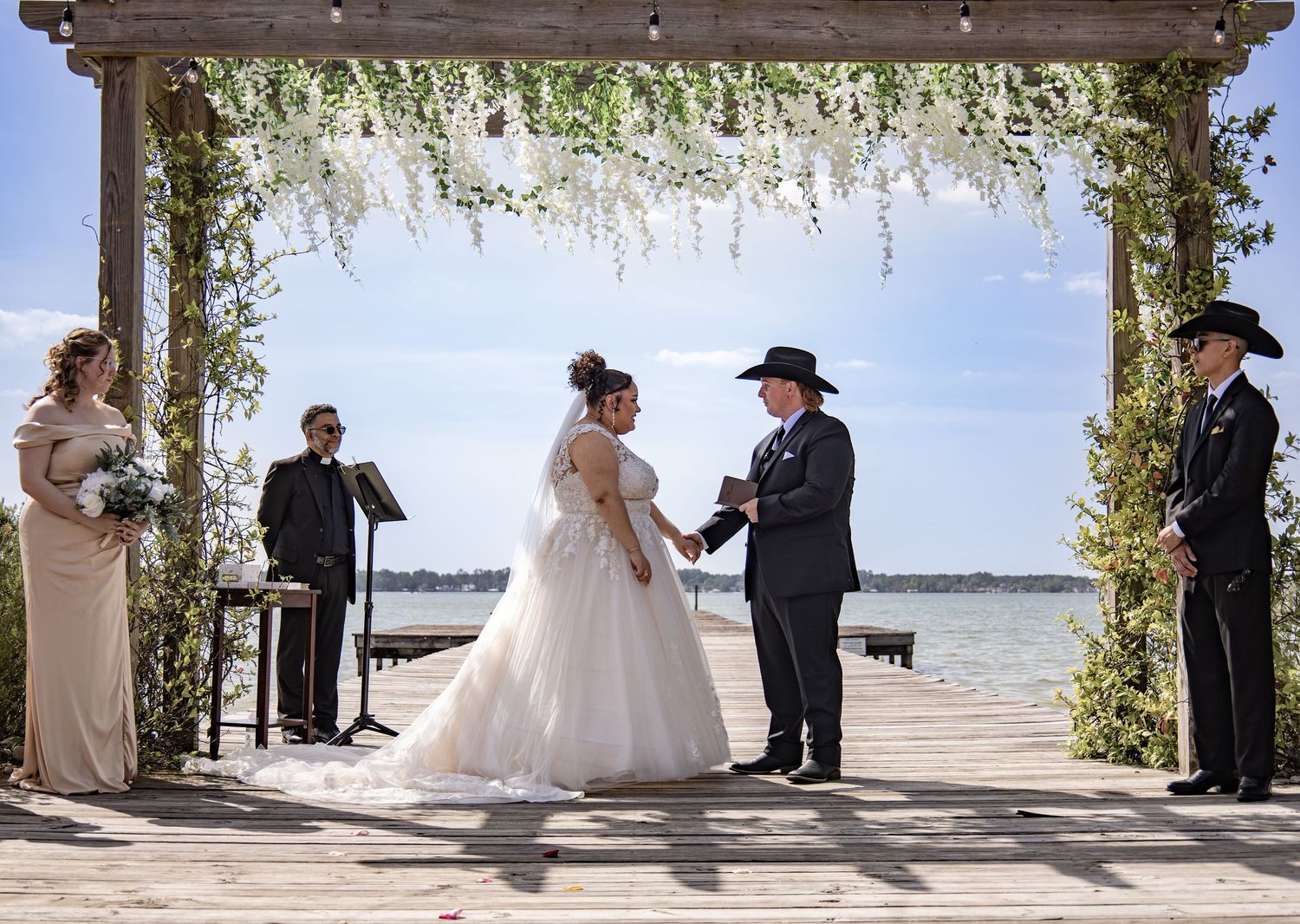 A wedding ceremony takes place under a floral-decorated pergola by the waterfront.